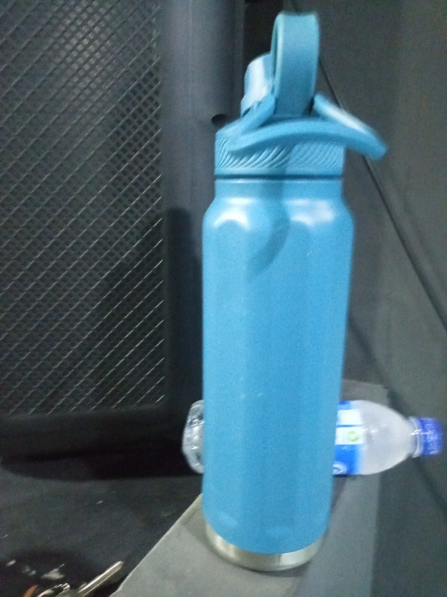 In the city of Juan Griego, Venezuela, a blue reusable water bottle stands upright on a dark gray surface. The bottle is tall and cylindrical, with a silver band around its base. It has a blue lid with a built-in carrying loop and a spout. To the right of the blue bottle, a partially visible clear plastic water bottle with a blue cap is present, lying on its side. The background consists of a textured dark gray surface on the left, which appears to be a grid pattern, and a smoother dark gray surface on the right.