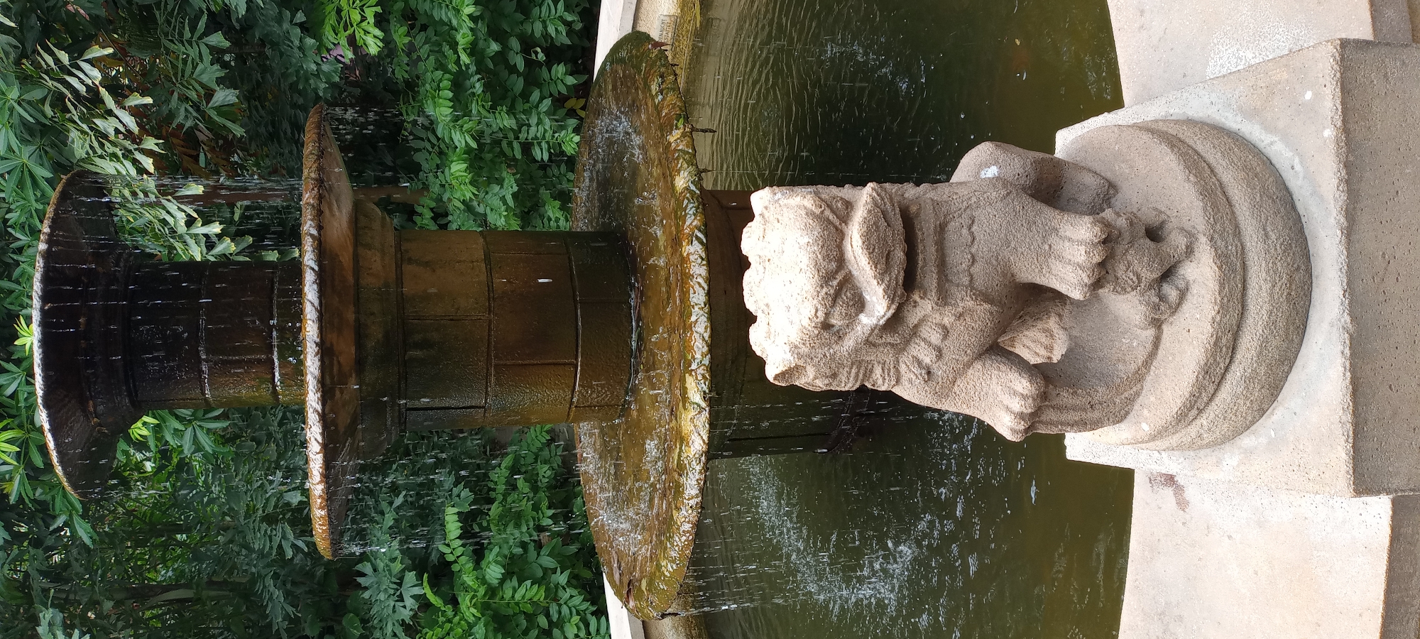 The image shows a stone lion statue in front of a multi-tiered fountain in what appears to be a garden setting. The fountain is made of dark stone and has water cascading down from its upper levels into a pool. Lush green foliage is visible in the background, suggesting an outdoor location. The lion statue is carved from a lighter-colored stone and sits on a circular base, with its paws resting on the edge of the pool. The statue has a stylized, somewhat fierce expression with its mouth open. The overall scene conveys a sense of tranquility and natural beauty. There are no people visible in the image. The location is not Gelsenkirchen, Germany, and there is no information available to identify the specific location in Gelsenkirchen.