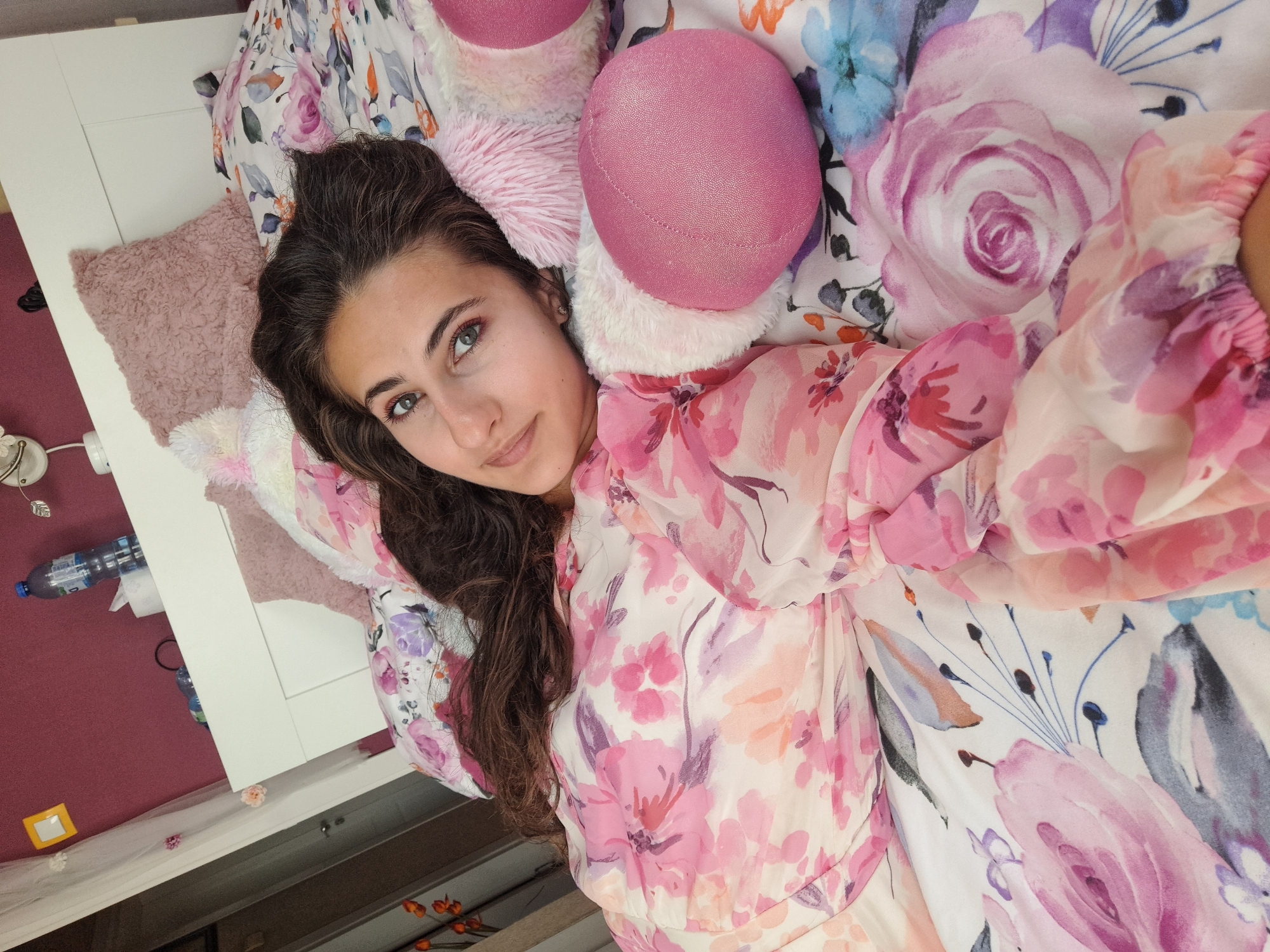 The media file is a selfie taken from above, showing a young woman lying on a bed. She has long, dark brown hair that is spread out around her. The woman is wearing a light, flowy pink dress with a floral pattern in shades of pink, purple, and blue. Her eyes are open and she is looking towards the camera with a slight smile.
The bed is covered with a duvet that has a similar floral pattern to her dress. There are several pillows on the bed, including a round pink one and a fluffy pink one. To the left of the bed, a white piece of furniture, possibly a dresser or nightstand, is visible. On this furniture, there are a couple of water bottles. The floor next to the bed is a dark pink color.
The lighting suggests it is daytime. The overall mood is relaxed and comfortable. The focus is on the woman and her surroundings, which are decorated with soft colors and floral motifs. There is no visible text in the media file.