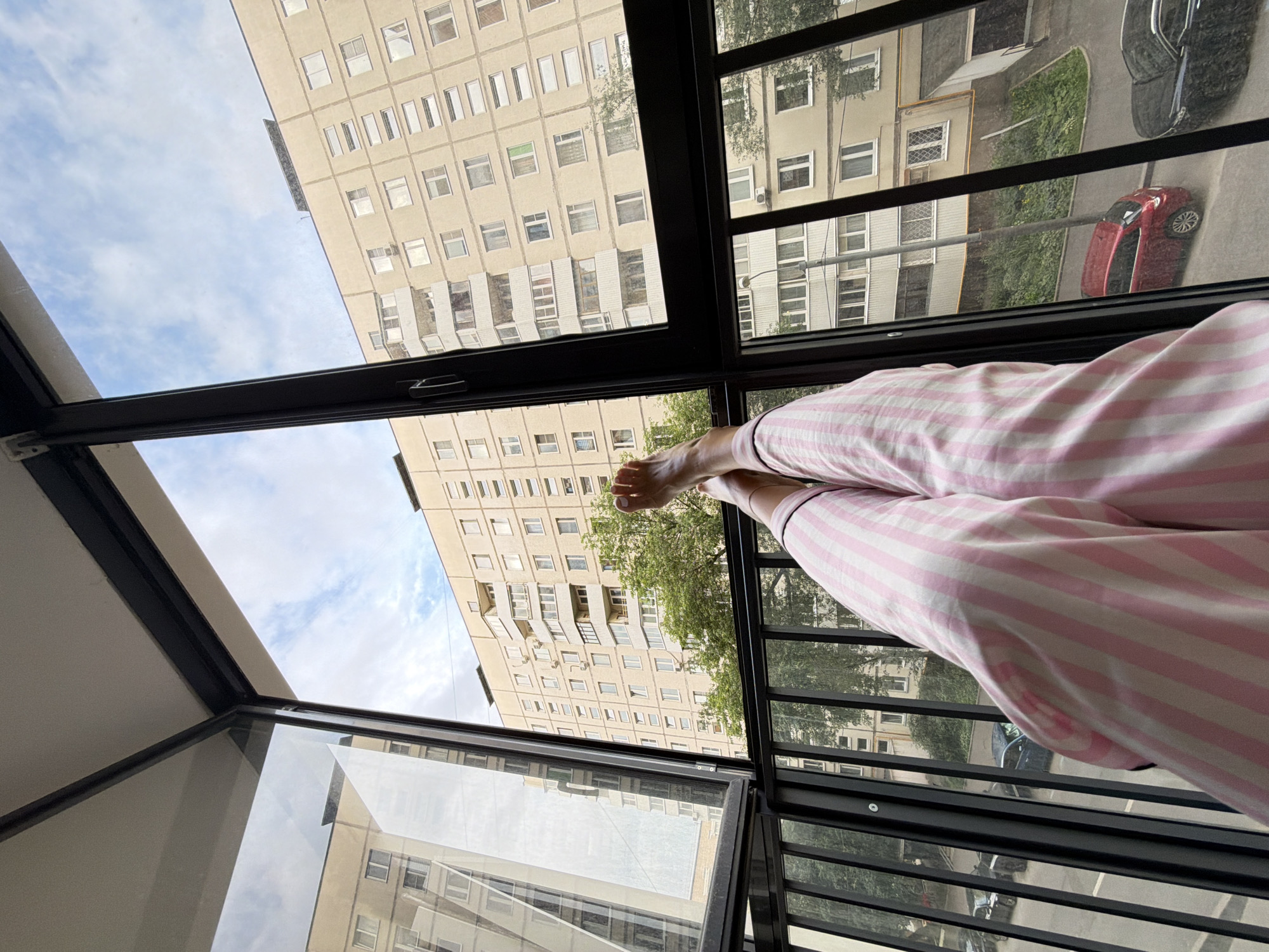 This media file captures a view from a balcony or window overlooking a residential area in Moscow, Russia. The perspective is from someone sitting with their legs extended, wearing pink and white striped pajamas. The subjects are the person's legs and feet, clad in pajamas, and the urban landscape outside.
The scene depicts a series of apartment buildings, typical of Soviet-era architecture, with many windows and balconies. Green trees are visible interspersed between the buildings, suggesting it is likely summer or early autumn. The sky is partly cloudy, indicating a daytime setting.
In the foreground, the glass enclosure of the balcony or window is apparent, reflecting the buildings and sky. Below, on the street level, a red car is parked. There are also reflections of other cars and what appears to be a dark-colored sedan. The overall impression is one of a relaxed, domestic moment, with the person enjoying the view from their home. The time of day is likely mid-morning to afternoon, given the lighting. No text is visible in the media.