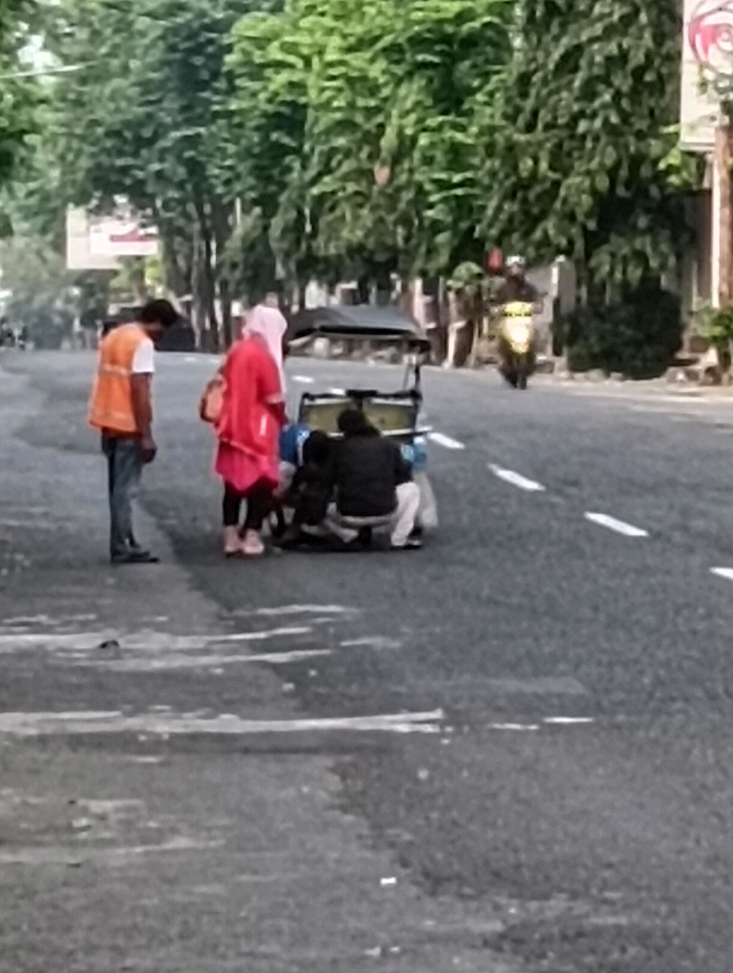 In the city of Purworejo, Indonesia, a street scene unfolds. The foreground features a paved road with visible white lane markings. A man in an orange vest and blue jeans stands on the left, looking down. Beside him, a woman in a pink hijab and pink top is crouched, attending to something on the ground, near a three-wheeled vehicle. Another person, wearing dark clothing and white pants, is also crouched next to the vehicle. In the middle ground, a motorcycle is being ridden by a person wearing a dark helmet and a yellow jacket, moving away from the viewer. The background is filled with lush green trees lining the street and buildings. The overall impression is of a moment of activity on a suburban street.