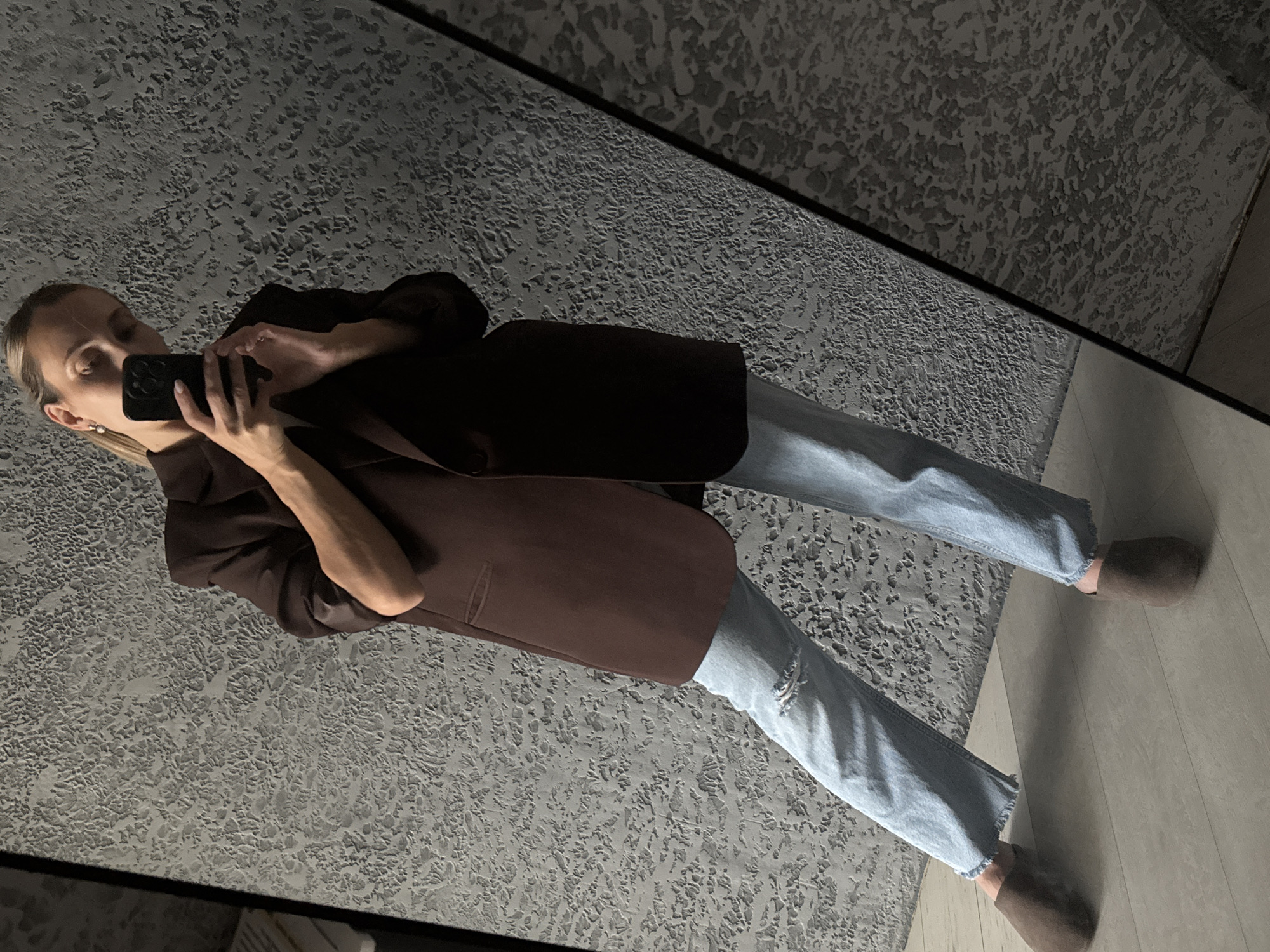 A woman stands indoors, taking a mirror selfie with her phone. She is wearing a dark brown oversized blazer, light blue ripped jeans, and dark brown slippers. The background consists of a textured grey wall and a light wood-grain floor. The image appears to be taken in a domestic setting. There is no information within the image that indicates the location is in the city of Chaikovsky, Russia.