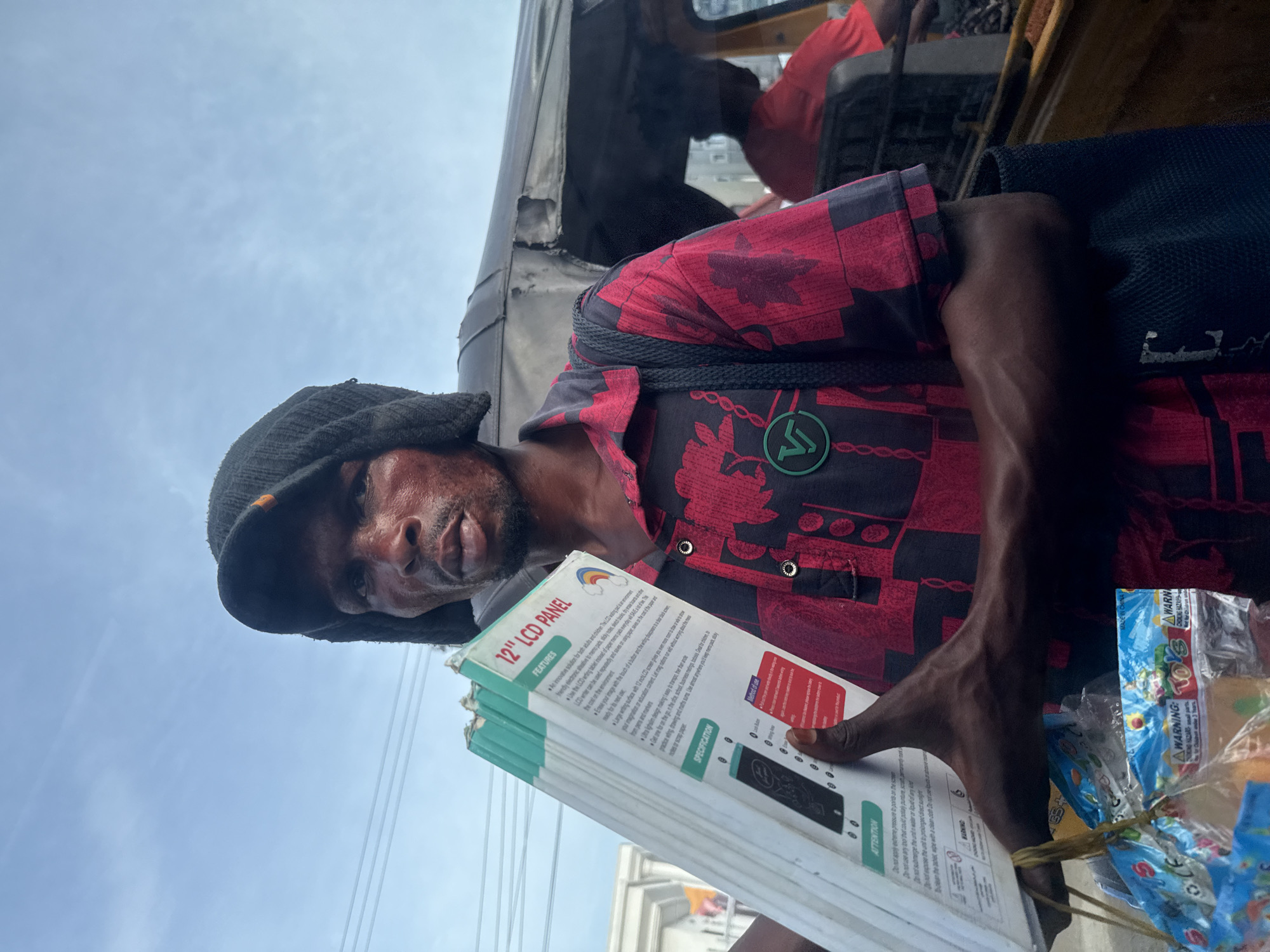 The image shows a man wearing a black beanie and a red and black patterned shirt. He's standing near a vehicle, possibly a tricycle taxi, and holding several boxes labeled "12" LCD PANEL". He is looking directly at the camera. In the background, another person can be seen seated inside the vehicle. Additionally, a package of toys is visible. The image was taken in Ado, Nigeria.