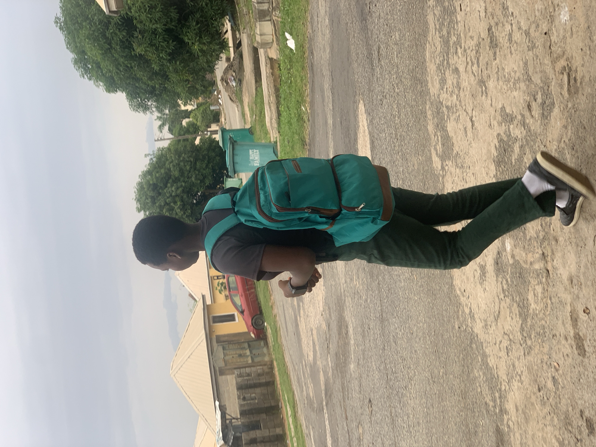 A person with dark skin and short hair, wearing a dark grey t-shirt and dark green trousers, walks from left to right across a paved road. They are carrying a large, teal backpack with brown accents. The person is looking away from the camera, towards the right side of the frame. Their right hand is slightly raised, and they appear to be wearing a watch on their left wrist.
The environment suggests a suburban or residential area. In the background, there are several yellow houses with beige roofs. A red car is parked on the side of the road. Green trees line the area behind the houses, and several large green trash bins are visible. The sky is overcast and grey. The road is paved but shows signs of wear and tear, with patches of lighter colored dirt and debris. The overall scene is consistent with a street in the city of Abuja, Nigeria.