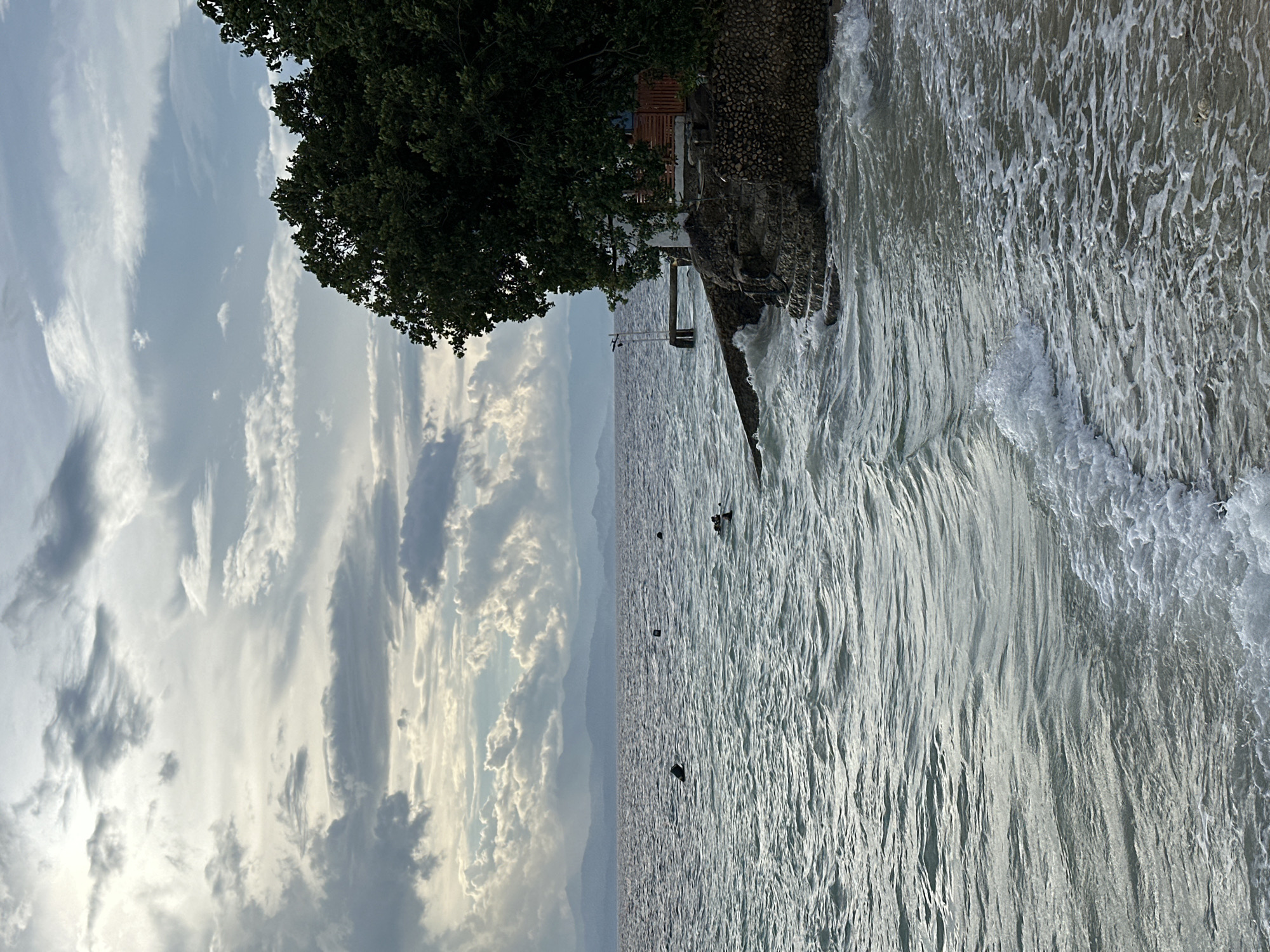 The image depicts a coastal scene on a cloudy day, likely in Basdiot, Philippines. The foreground features the choppy, gray surface of the ocean with white foam from small waves lapping onto a sandy shore. The water is a mid-tone gray with highlights reflecting the overcast sky.
In the mid-ground, the ocean extends towards a distant, hazy coastline marked by layered mountains. Several dark, cylindrical objects, possibly buoys or markers, are visible scattered across the water. A few figures, appearing small in the distance, are in the water, suggesting recreational activity like swimming or snorkeling.
To the right, a rocky, stone-lined embankment rises from the water, topped with lush green trees. A small wooden structure, possibly a hut or dwelling with a wooden fence and a small dock extending into the water, is situated on this elevated land. The architecture of the structure appears simple and rustic. The trees overhang the structure and the embankment, creating a dense canopy.
The sky is filled with a mix of dark gray and lighter, wispy clouds, indicating overcast or potentially stormy weather. The overall impression is one of a natural, somewhat wild coastal environment, with signs of human presence in the form of the structures and the buoys.