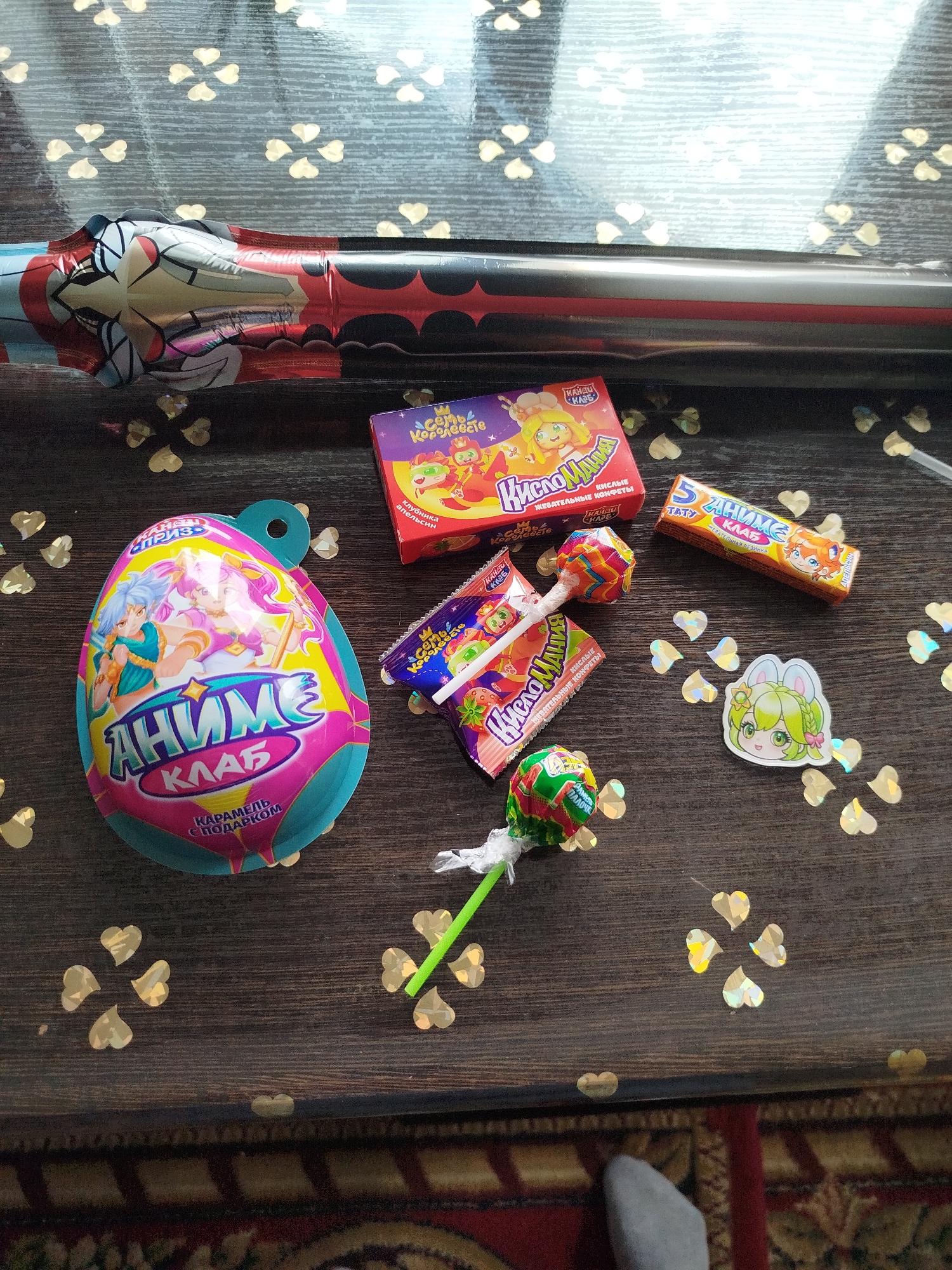This is a top-down, close-up shot of various confectionery items and a foil balloon scattered on a dark wooden surface. The scene appears to be an indoor setting, possibly a table, with a patterned rug visible at the bottom of the frame. Golden, heart-shaped confetti is sprinkled across the surface, adding a festive touch.
The subjects include an oversized, egg-shaped package with an anime-style illustration of two characters and the text "АНИМЕ КЛАБ" (Anime Club) and "КАРАМЕЛЬ С ПОДАРКОМ" (Caramel with a gift). Beside it is a small red box labeled "семь Королевств" (Seven Kingdoms) with cartoon characters and the text "клубника апельсин" (strawberry orange). There are also two individually wrapped candies with illustrations and text including "КИСЛЫЕ ЖЕВАТЕЛЬНЫЕ КОНФЕТЫ" (Sour chewy candies) and "КислоМания" (Sour Mania). A yellow wrapper with "5 АНИМЕ КЛАБ" (5 Anime Club) and "ЖЕВАТЕЛЬНАЯ РЕЗИНКА" (Chewing gum) is also present. Two lollipops are visible, one with a striped wrapper and another with a green wrapper, partially obscured by confetti. A small sticker of an anime character with bunny ears is also on the surface.
A long, metallic foil balloon, decorated with a comic-book style illustration, runs horizontally across the top of the image. The overall impression is that of a collection of children's candy and party favors, likely presented as a gift or a collection of treats. There are no specific cues for time of day or weather. The text is primarily in Russian, indicating a possible location in or related to Russia or a Russian-speaking region.