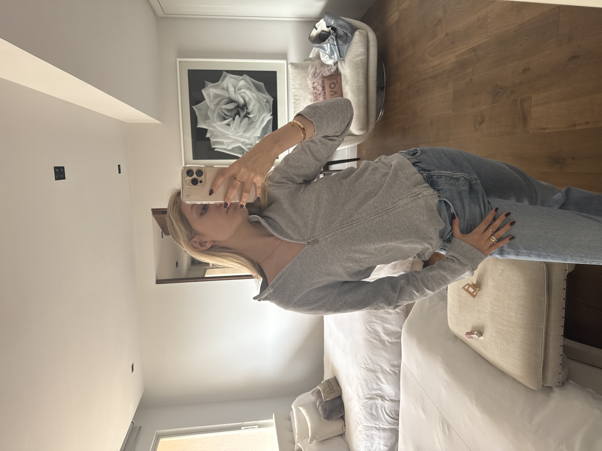 A person is taking a selfie in what appears to be a bedroom. The room features white walls and light-colored wooden flooring. The bedroom contains a bed with white bedding, a cushioned headboard, and several pillows. There is a chair in the background, along with a framed picture on the wall. The person in the selfie is wearing a gray zip-up jacket and jeans, and is holding a smartphone. It's not possible to determine the location in Mexico City from the image alone.
