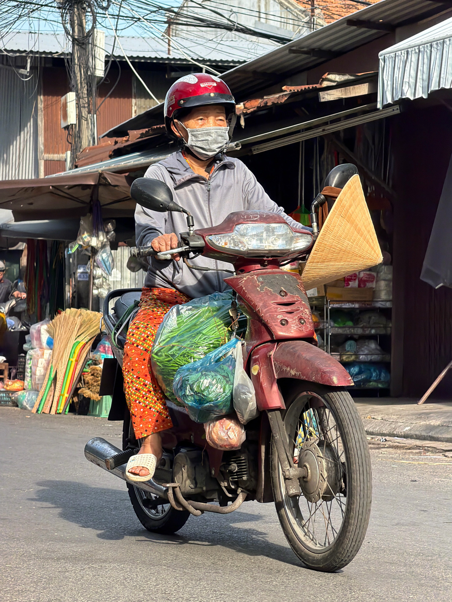 An elderly person, wearing a red helmet with a black stripe and a grey medical mask, rides a red motorcycle through a street in Nha Trang, Vietnam. They are dressed in a grey jacket, an orange and yellow patterned skirt, and white sandals. The motorcycle is laden with bags of fresh produce, including green vegetables, and a conical hat is attached to the handlebars. In the background, there are stalls with goods for sale, including brooms, and a tangle of overhead wires. The scene appears to be a market street, with buildings and awnings lining the road. The sunlight suggests it is daytime.