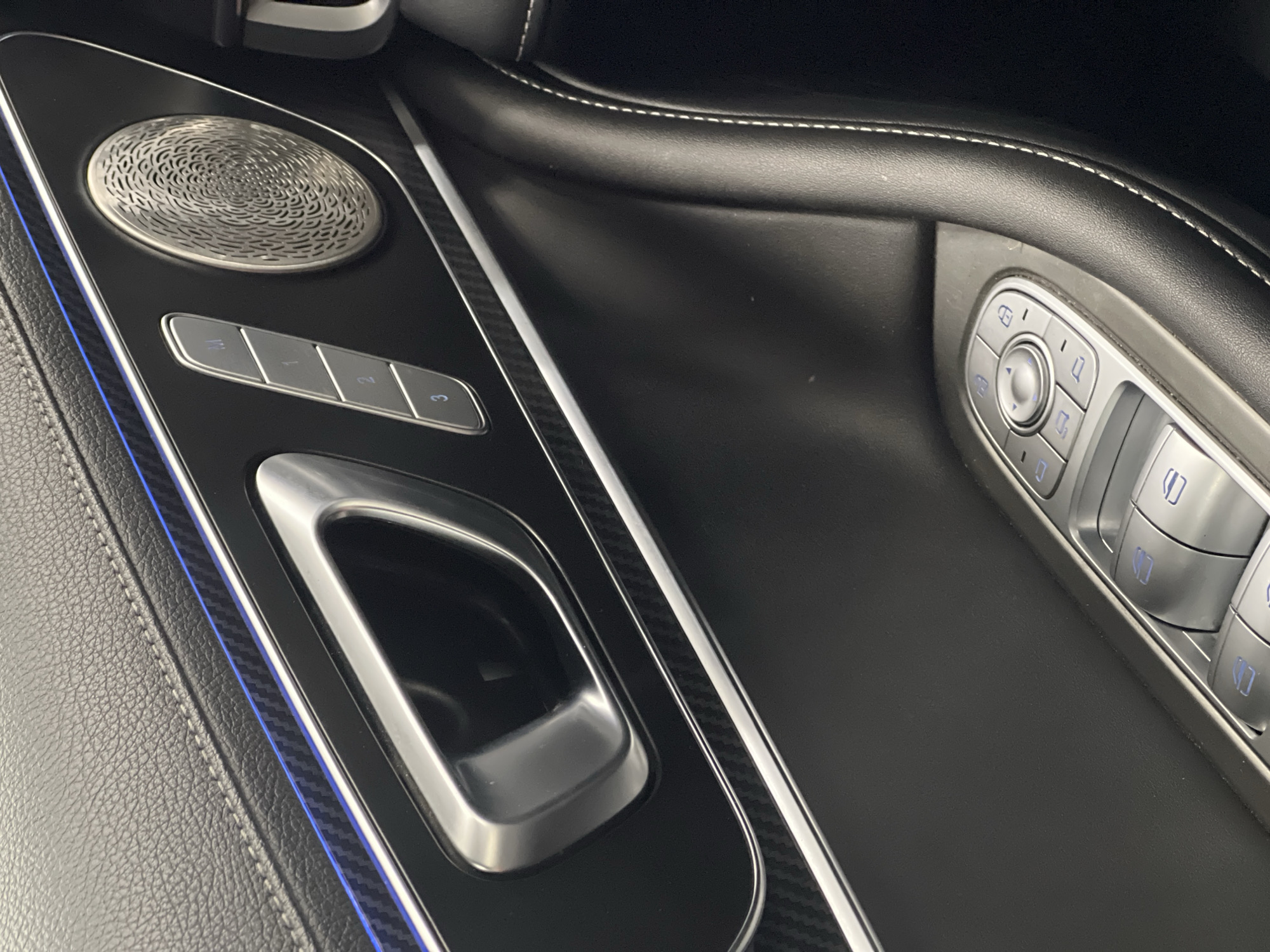 The image presents a detailed close-up of the interior front door panel of a luxury vehicle. The primary material is black, finely textured leather with contrasting white stitching visible along the top edge and vertically on the right side of the frame. A horizontal trim piece extends across the mid-section of the panel, featuring a dark carbon fiber-like pattern bordered by thin, polished metallic strips. A narrow blue LED ambient light strip is illuminated just above the carbon fiber trim.
To the left, a polished metallic door handle is recessed into the panel. To the right of the door handle, a control panel with four elongated silver buttons is arranged vertically. These buttons are labeled 'M', '1', '2', and '3', indicating memory seat settings, and show faint blue illumination. Adjacent to these buttons is a circular, polished metallic speaker grille with an intricate, swirling geometric pattern.
In the lower section of the door panel, a cluster of window and mirror controls is visible. These are silver-colored, ergonomically shaped buttons with blue illuminated icons. They include a circular joystick-like control for mirror adjustments, surrounded by four smaller buttons (likely for mirror folding and selection), and four rocker-style switches for power windows. Two lock/unlock buttons are also visible within this cluster.
The overall environment suggests a well-maintained, modern vehicle interior with emphasis on refined materials and technology. The image contains no visual information identifying its geographic location as Izhevsk, Russia, or any other specific city.