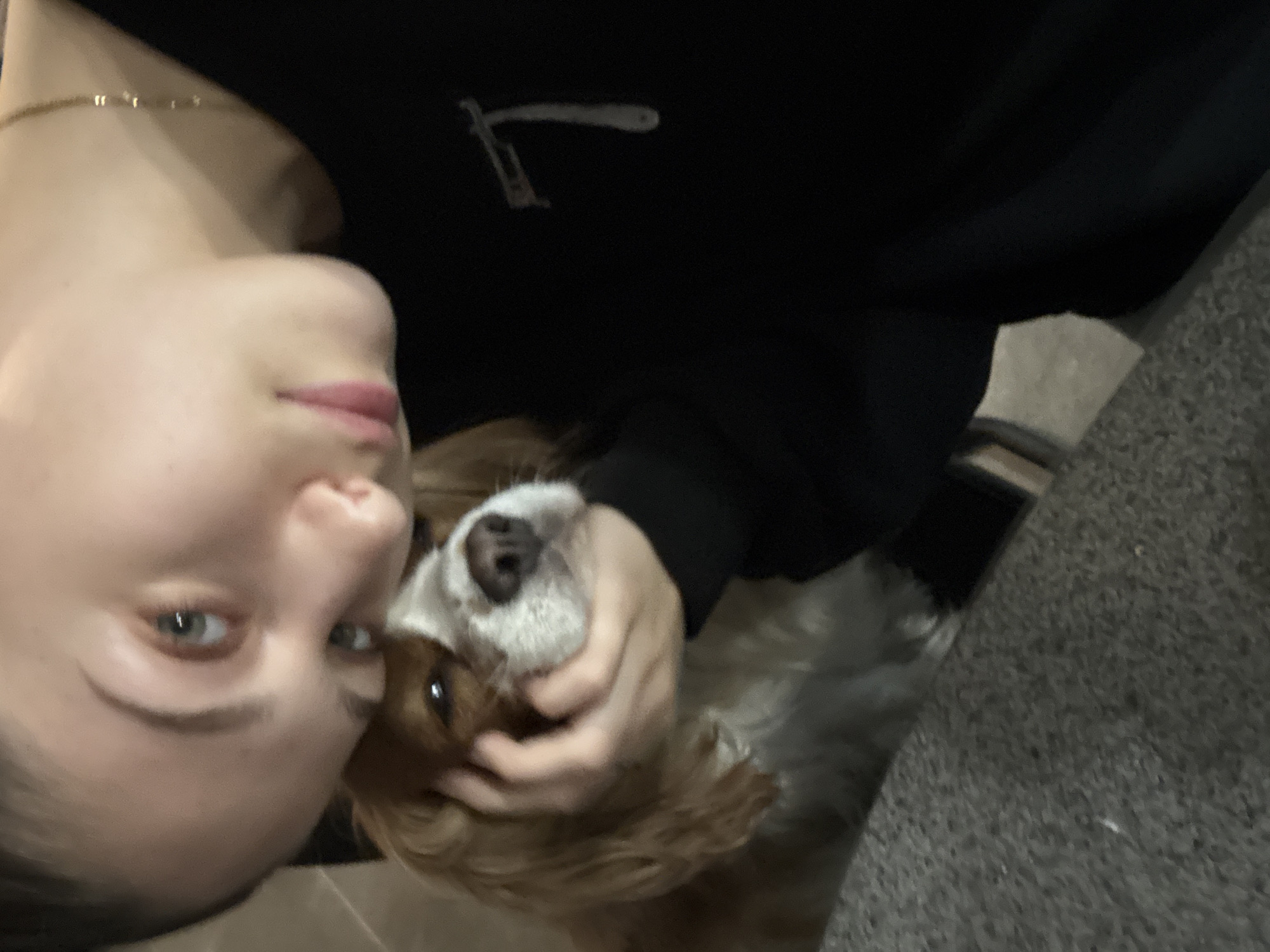 This image depicts a young adult female in an indoor setting in Sofia, Bulgaria, engaged in close physical interaction with a dog. The female, positioned on the right side of the frame with her head tilted, has light-colored eyes directly engaging the viewer, fair skin, and dark hair pulled back. She is wearing a black long-sleeved garment with a faint, light-colored, stylized 'T' or abstract symbol visible on the upper left chest area, and a thin gold chain around her neck. Her left hand is gently cradling the head and muzzle of a small, long-haired dog, which appears to be a King Charles Spaniel or similar breed. The dog, positioned to the lower left, exhibits tan/brown and white fur, with dark eyes and a dark nose, looking slightly away from the camera. The female's face is positioned close to the dog's head, appearing to rest gently upon it. The foreground features a grey, textured surface, possibly carpet or upholstery. The background is dark and out of focus, providing no specific environmental details beyond an internal space.