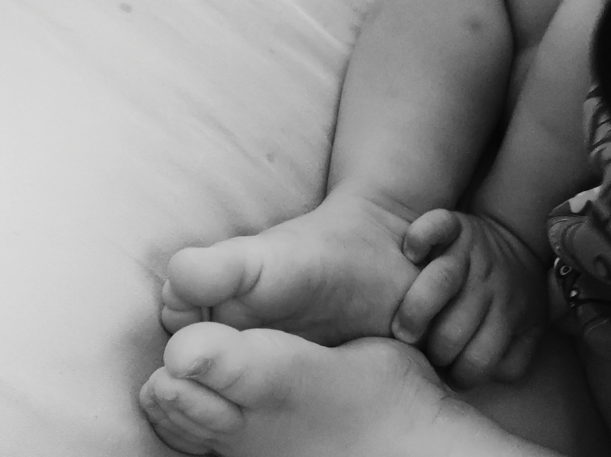 A black and white, close-up photograph shows the feet and hand of a child. The feet are resting on what appears to be a light-colored, possibly textured surface. The right hand of the child is grasping the toes of the left foot. The image is cropped, so only a portion of the child's legs and torso are visible on the right side of the frame. No information in the image suggests a specific location, such as the city of Chaikovsky, Russia.