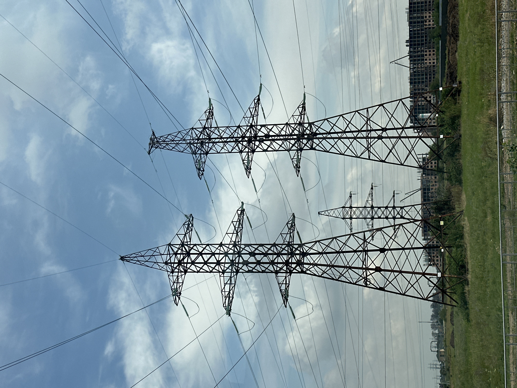 In Beryozovy, Russia, the image captures a panoramic view of a landscape dominated by towering electricity pylons. Three large pylons stand prominently in the foreground, their lattice structures silhouetted against a cloudy blue sky. The pylons are strung with thick cables, stretching across the frame and disappearing into the distance. Beyond the pylons, a vast expanse of green grass stretches out, dotted with bushes and small trees. In the middle ground, the skeletal frames of several multi-story buildings under construction are visible, indicating ongoing development. A few smaller electricity pylons and industrial structures can be seen in the far distance, hinting at a larger industrial or infrastructural complex. The overall scene conveys a sense of industrialization and development in a semi-rural setting.