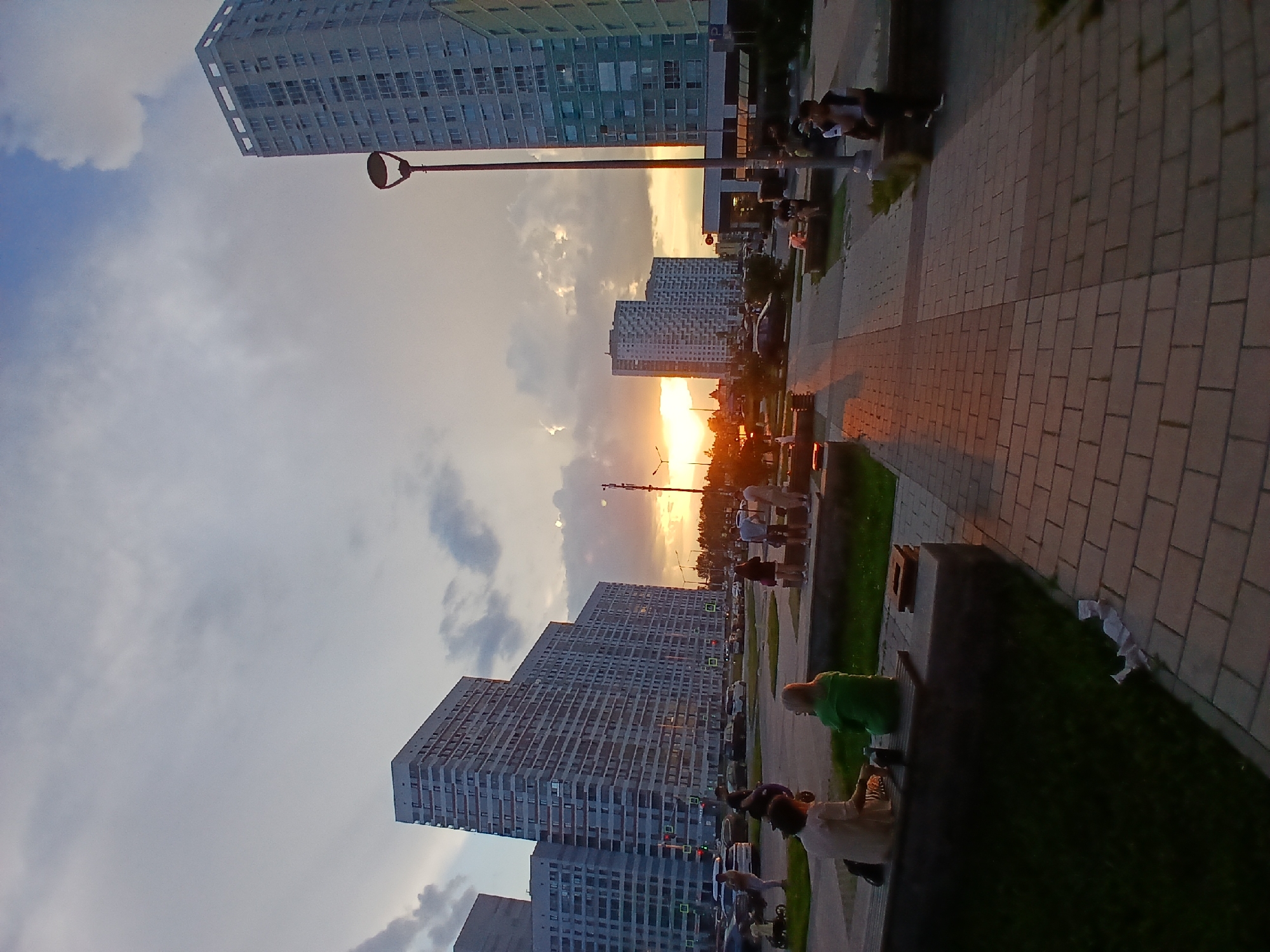 The image captures a sunset scene in a modern urban environment, likely in Yekaterinburg, Russia, characterized by high-rise apartment buildings. In the foreground, a paved walkway leads toward a park area with benches. Several individuals are seated on the benches, some facing away from the camera, engaged in conversation or resting. A person in a green jacket is visible on a bench, alongside another individual wearing a light-colored shirt. Further into the park, more people are scattered on benches and standing.
Dominating the background are several tall, multi-story buildings with numerous windows, indicative of residential complexes. The setting sun casts a warm glow, illuminating the sky with hues of orange and yellow, and creating long shadows across the paved areas. The presence of streetlights suggests that this is a public space designed for residents. Cars are parked and driving on a street visible behind the park area, implying a busy urban setting. The overall atmosphere is one of a calm, late afternoon or early evening gathering in a developed residential district.