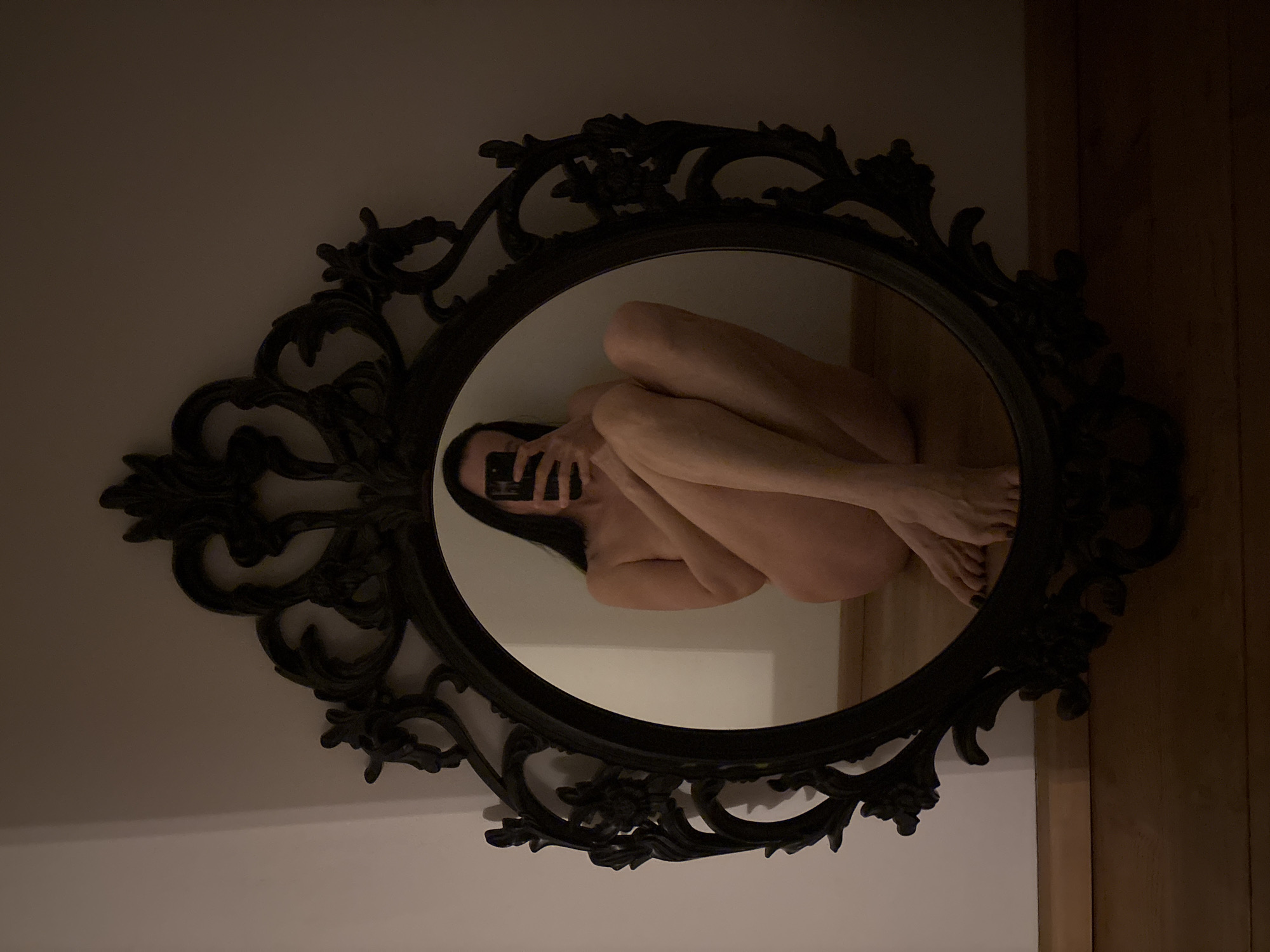 A selfie of a nude person is reflected in a mirror with an ornate, oval, black frame. The person is seated on the floor with their knees drawn up to their chest, holding a black smartphone in front of their face. The mirror is mounted on a light-colored wall next to a wood-paneled wall. The location in the city of Chaikovsky, Russia, is undetermined.