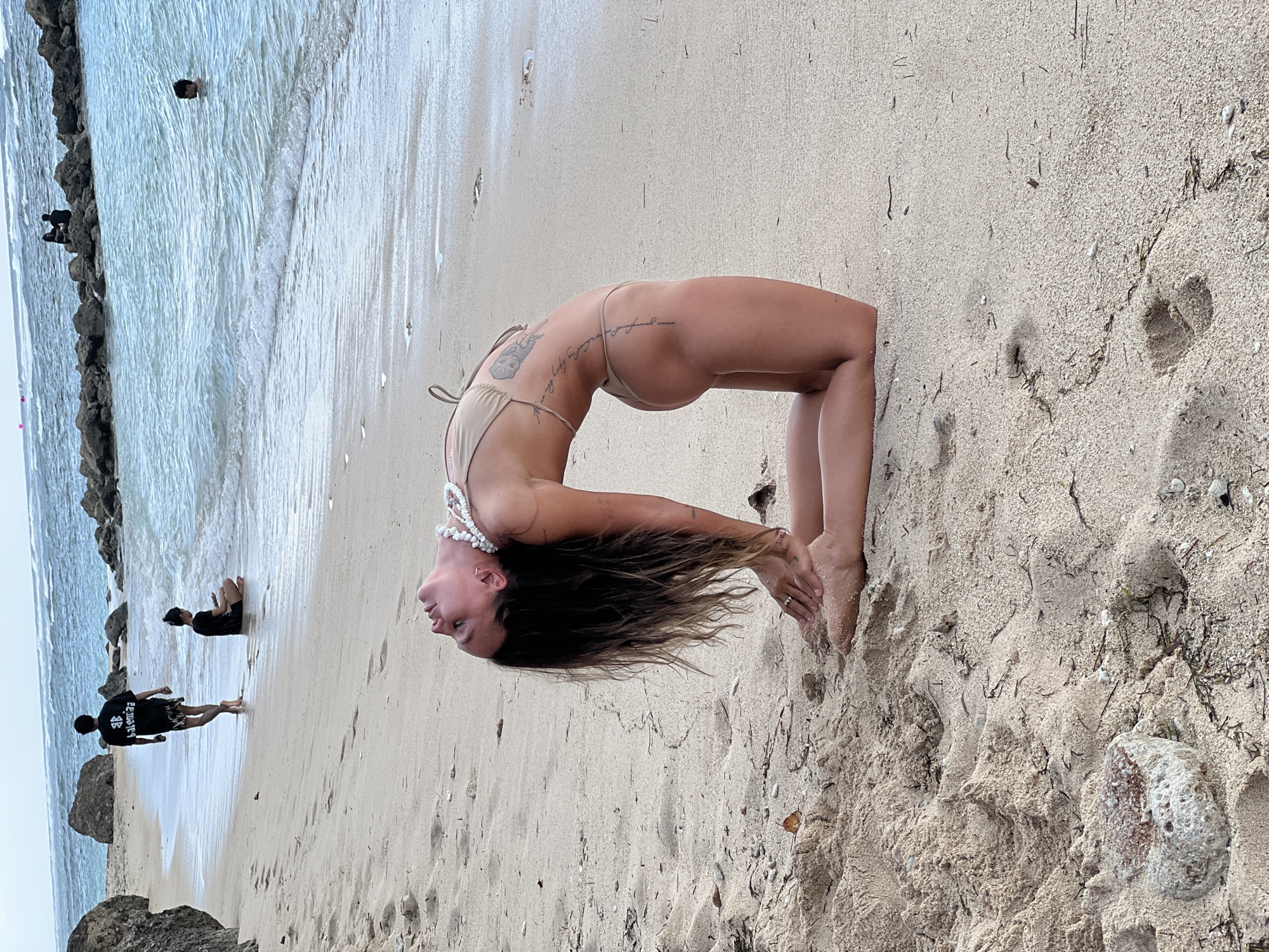 A woman in a light-colored bikini is performing a yoga backbend on a sandy beach. The woman's knees are on the sand, and her torso and head are bent backward so that her head is facing the ground, and her hands are reaching for her feet. She has long dark hair that flows towards the ground and is wearing a white necklace. In the background, there is a group of people near the water, and some rocks or a stone barrier are along the shoreline. The setting is likely a beach in Kuta Selatan, Indonesia, based on the environment and coastal characteristics.