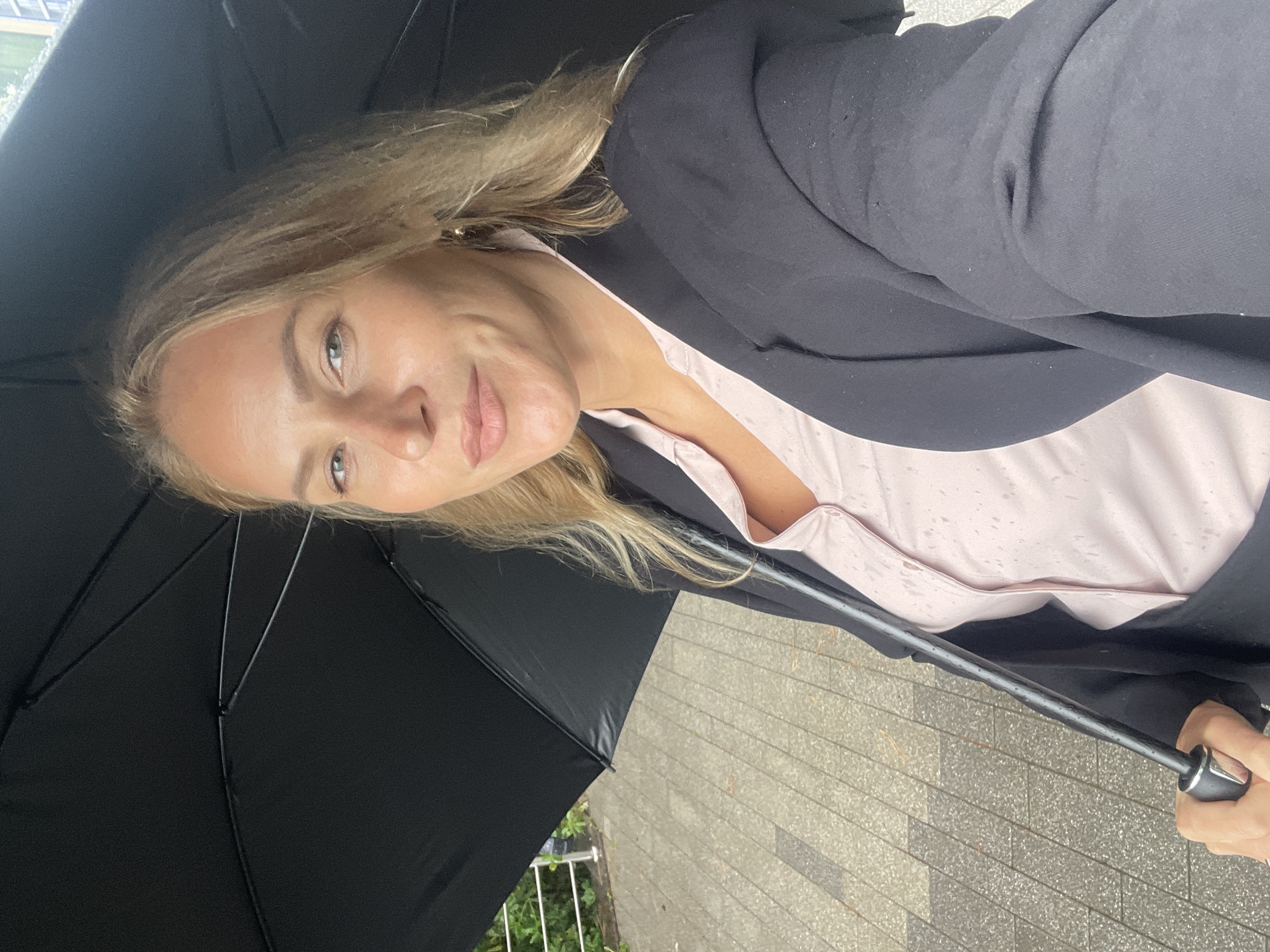 A woman is holding a black umbrella outdoors. She is wearing a black blazer and a light pink shirt. The ground appears to be paved, possibly in a city setting. There is a partial view of greenery and a railing. The image does not confirm the city of Sochi, Russia.