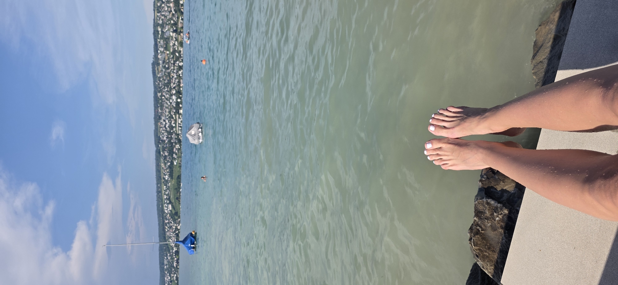 In Oberrieden, Switzerland, a first-person perspective shot shows a person's bare legs and feet dangling over a stone embankment and into a body of water. The feet are clean with white nail polish. The water is calm and has a greenish hue, with ripples on the surface. In the background, a town with white buildings is visible on the shore, nestled against green hills under a blue sky with scattered clouds. A sailboat with a blue sail and another covered boat are anchored in the water. A few people are swimming in the distance. The overall scene depicts a sunny day at a lake or large body of water.