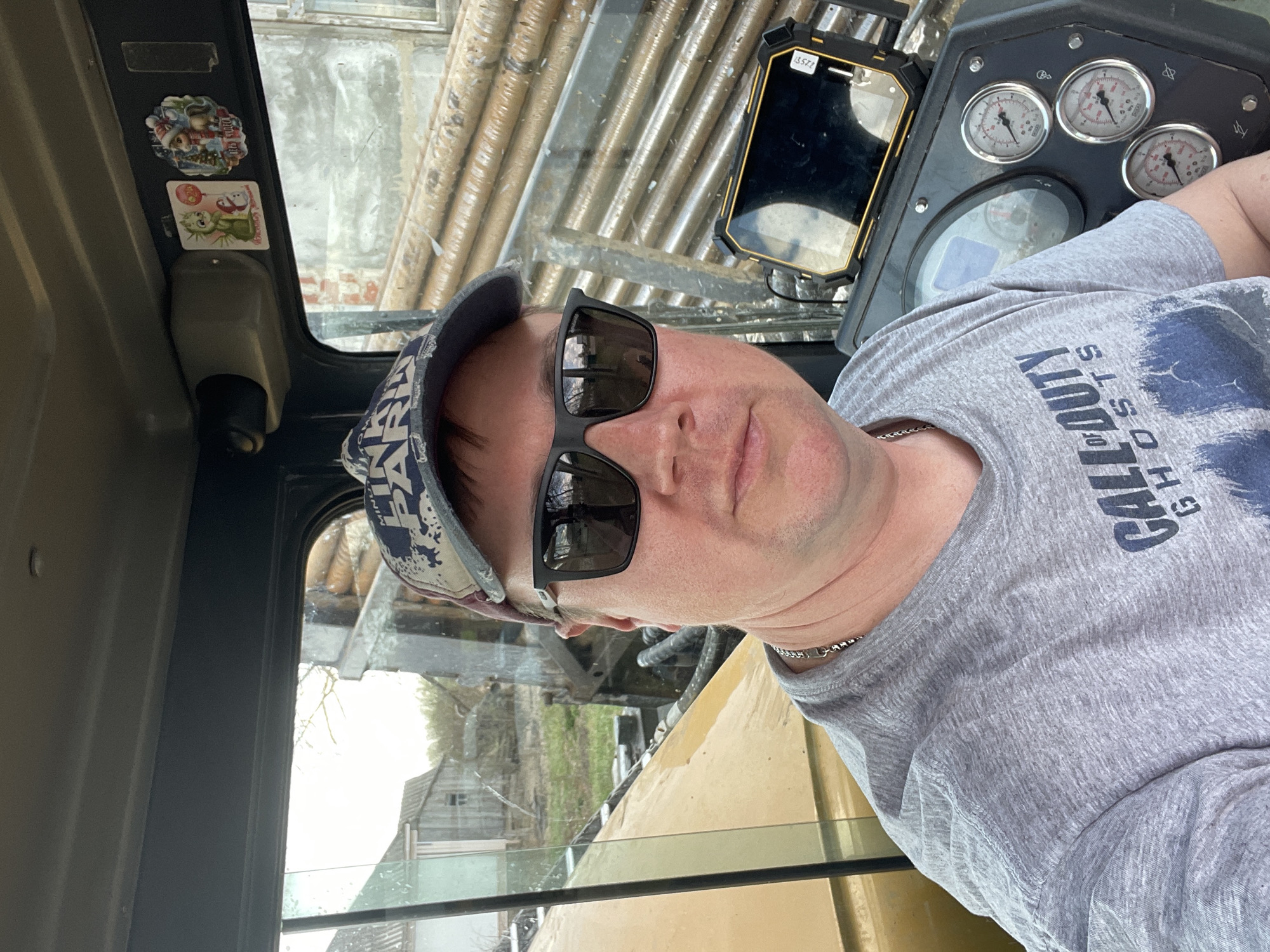 The image shows a man wearing sunglasses and a camouflage hat inside the cabin of heavy machinery. He is wearing a gray t-shirt with "Call of Duty Ghosts" printed on it and a silver chain. The machinery includes gauges, a control panel, and a rearview mirror with a cell phone attached to it. Outside the cabin, one can see a building, possibly a house, through the glass and metal bars. Based on the image’s geotag, the scene is located in Поедуги, Russia.