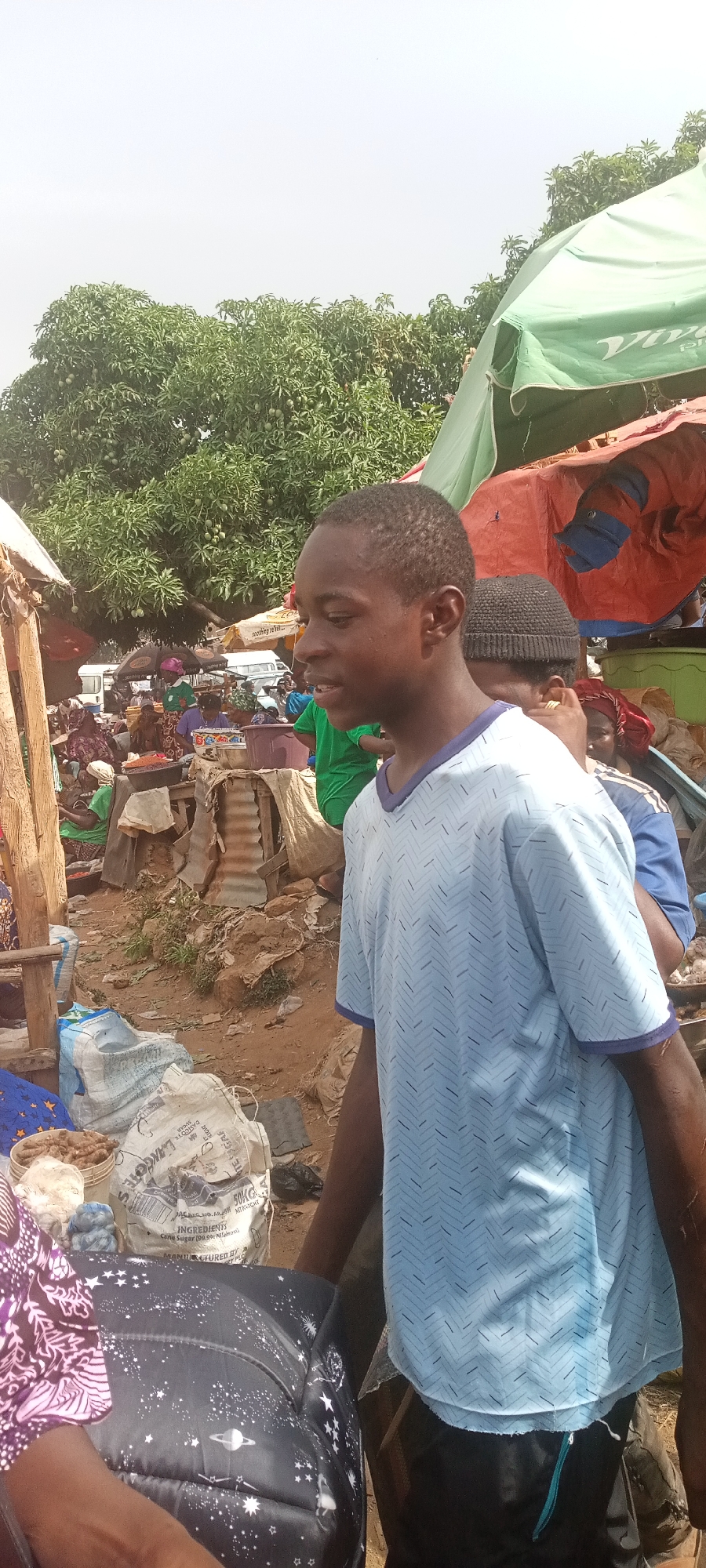 The image shows a young Black African male wearing a light blue, short-sleeved V-neck shirt with a repeating pattern, standing in an open-air market in Gura Top, Nigeria. The market is bustling with people and activity, with stalls covered by assorted canopies and umbrellas. Behind the subject, there is another person wearing a black knitted hat and a blue shirt.
The market setting is somewhat crowded and disorderly, with items like bags of sugar from "DANCOTE" visible on the ground, suggesting the presence of vendors and shoppers. The ground appears to be dirt or mud. There are other people engaged in buying and selling transactions. In the background are trees, more stalls, and a white van. The sky is overcast, indicating a cloudy day.