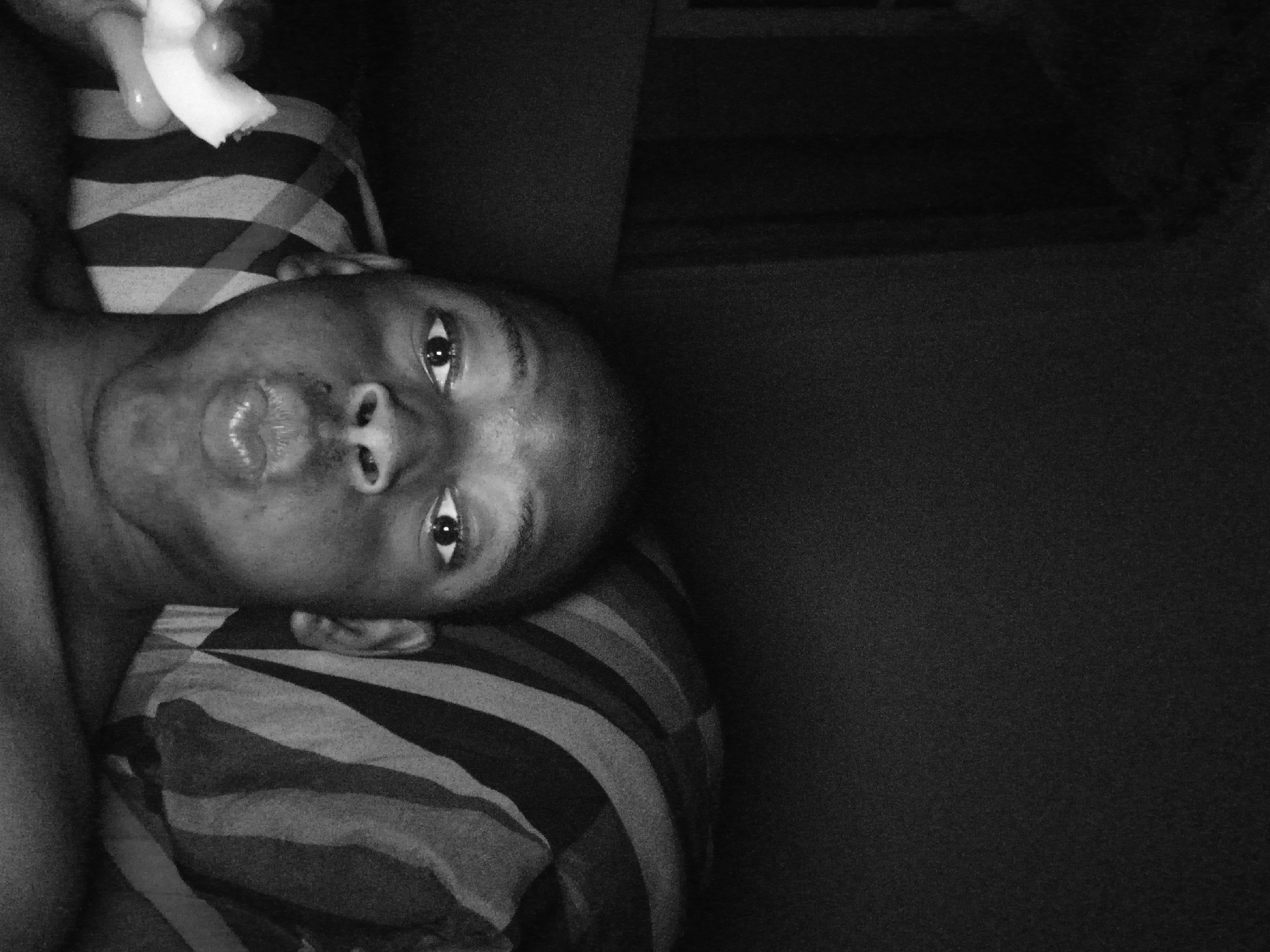 A black and white photograph depicts a person lying down, possibly on a striped pillow. The person is light-skinned and their lips are pursed. In the upper left corner of the image is someone's hand holding an object. The rest of the image shows the darkness of the room.
I cannot identify the location as Lagos, Nigeria.