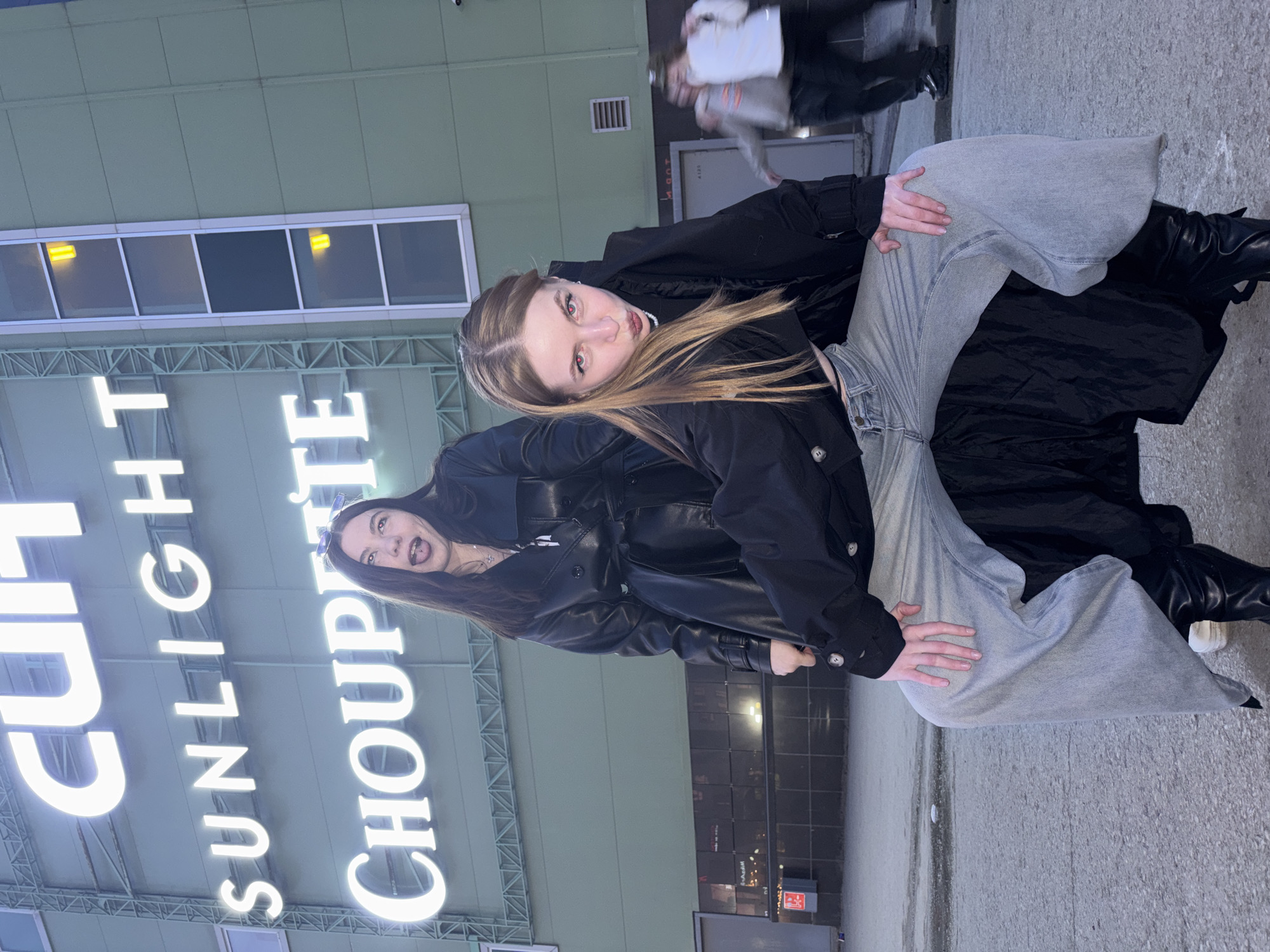 Two women are posing for a picture in Perm, Russia, in front of a building with the words "CULT SUNLIGHT CHOUPETTE" illuminated in white neon letters. One woman is wearing a black jacket and light gray jeans, and she has dark blonde hair. She is holding the other woman up by her legs. The other woman is wearing a black leather jacket and has dark hair. Both women are smiling. The background includes the building facade, windows, and a blurred figure walking by. The ground is concrete.