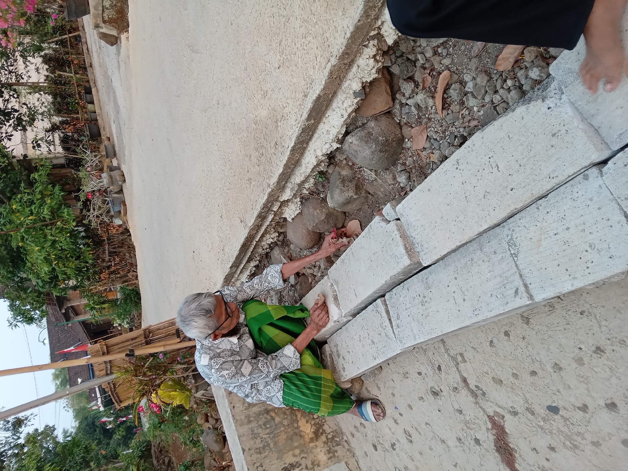 In Kedunggudel, Indonesia, an elderly person with gray hair and glasses kneels on a concrete surface next to a row of concrete blocks. They are wearing a patterned shirt and a green sarong. The person is holding a small object, possibly a tool or material, and appears to be working on or interacting with the concrete blocks. The environment includes a dirt path with scattered stones and dried leaves, and vegetation including trees and potted plants in the background. A road and a building are also visible. The overall scene suggests an outdoor setting, possibly a residential area, with the person engaged in some form of manual activity.