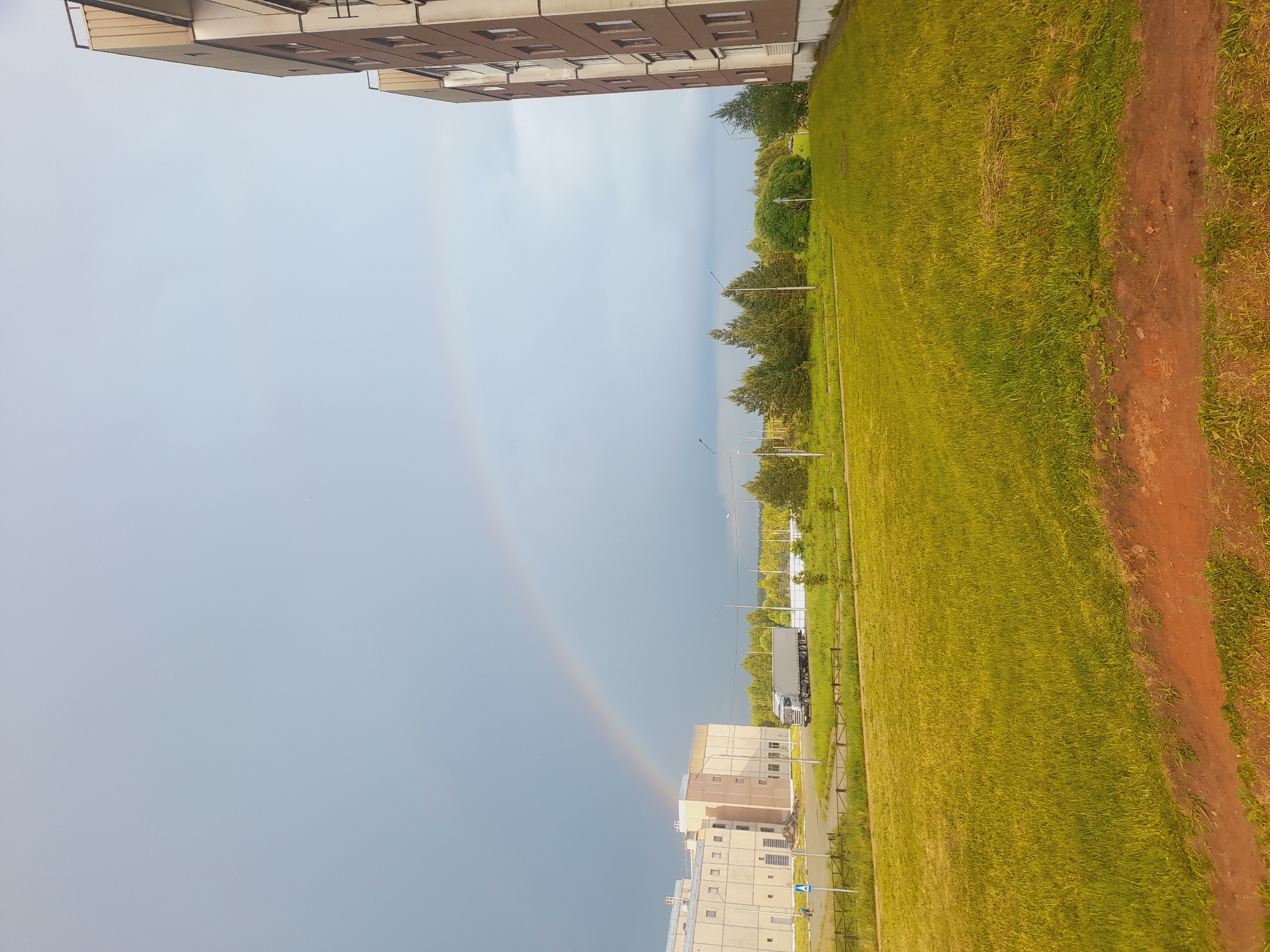 The image shows a rainbow arching across the sky. Below the rainbow is a building, and to the right of the building is a large grassy field. A truck is parked next to the field. In the upper portion of the image, there is a building and trees. The location appears to be a city with residential buildings and green spaces.