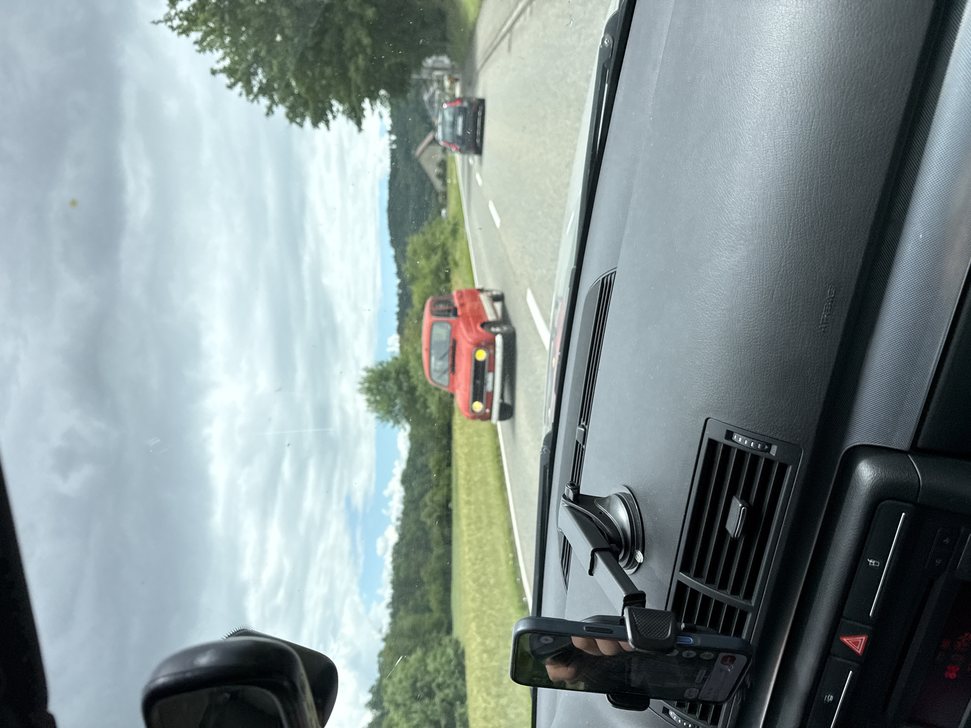 The image depicts a daytime road scene in a rural or semi-rural area, viewed from inside a car in Zurzach, Switzerland. The sky is bright but cloudy, overlooking expansive green fields, dense trees, and distant hills. On the asphalt road ahead, marked with white lines, a distinctive red vintage car (possibly a Renault 4) is directly in front, followed by a darker modern vehicle further down the road. A building is faintly visible behind the second car amidst the trees.
The foreground shows the interior of the car, featuring a dark grey, textured dashboard. An air vent is present on the right, and a phone holder, secured by a suction cup, clutches a dark smartphone. A hand is visible holding the phone. A rear-view mirror is partially seen on the left. The dashboard prominently displays an "AIRBAG" emblem on the passenger side. A control panel to the right includes a red triangular hazard warning button. The scene portrays a calm journey through scenic countryside under an overcast sky.