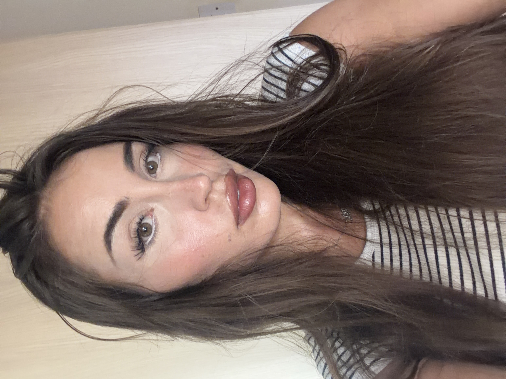 A medium shot shows a young Caucasian woman from the chest up, looking directly at the camera. She has long, dark brown hair that falls past her shoulders. Her skin is fair with rosy cheeks. She is wearing makeup, including white eyeliner on her lower waterline, dark mascara, defined eyebrows, and glossy pink lipstick. She is wearing a white and black striped t-shirt and a silver necklace. The background is a plain, light-colored wall with some vertical paneling. The lighting appears to be natural and coming from the right side of the frame. The image is a selfie taken indoors. The location is not specified in the image itself.