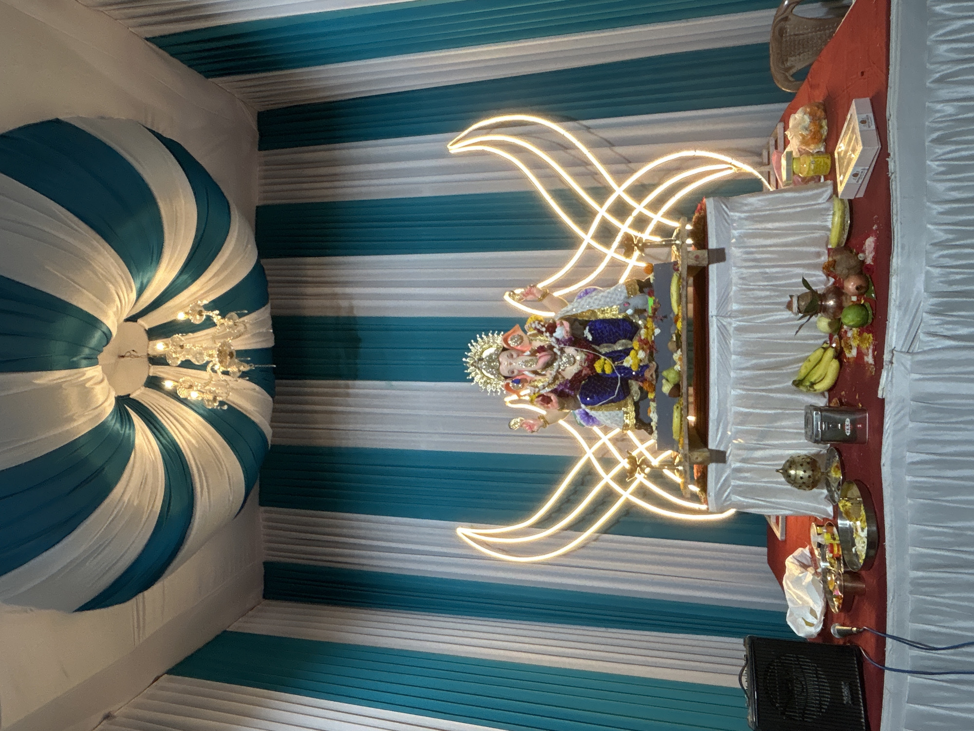 A richly decorated altar dedicated to Lord Ganesha is set up indoors. The altar is adorned with white and teal fabric drapery, creating a striped pattern on the walls and a swirled canopy overhead, from which a chandelier hangs. The central figure is a statue of Lord Ganesha, richly adorned and seated on a raised platform. Behind the statue, illuminated golden light fixtures form an abstract, wing-like design.
In front of the altar, a red-clothed table is laden with various offerings and items. These include plates of food, bananas, pomegranates, other fruits, and what appear to be sweets or Prasad in boxes. A speaker and microphone are positioned on the left side of the table, suggesting the setup might be for a religious ceremony or celebration with music. The overall environment appears to be a temporary setup, possibly for Ganesh Chaturthi celebrations, in the city of Mira-Bhayander, India. The scene is well-lit, highlighting the festive decorations and the deity.