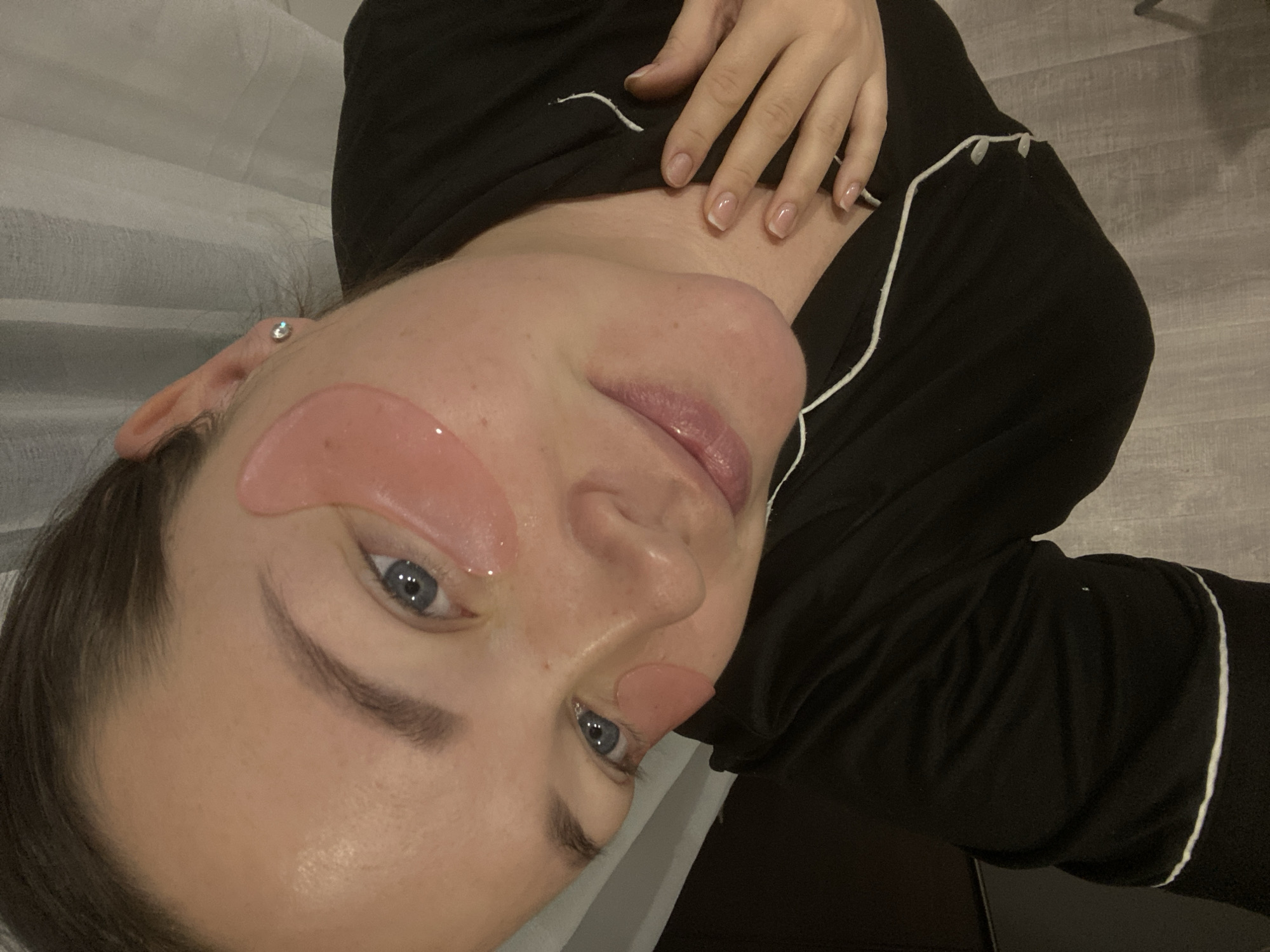This image depicts a close-up portrait of a light-skinned individual, presumably female, situated in an indoor environment, likely a private residence or hotel room in Sochi, Russia. The individual has dark hair, pulled back, and blue eyes. Two translucent pink hydrogel eye masks are applied beneath each eye. A small, clear stud earring is visible in her right earlobe. She is wearing a black garment with white piping, consistent with pajamas or loungewear. Her lips appear glossy, and her fingernails display a French manicure. Her left hand rests gently on her upper chest, below the collarbone. The background includes light-colored curtains and a light grey floor with a wood-effect pattern. The individual is looking towards the viewer with a subtle smile.