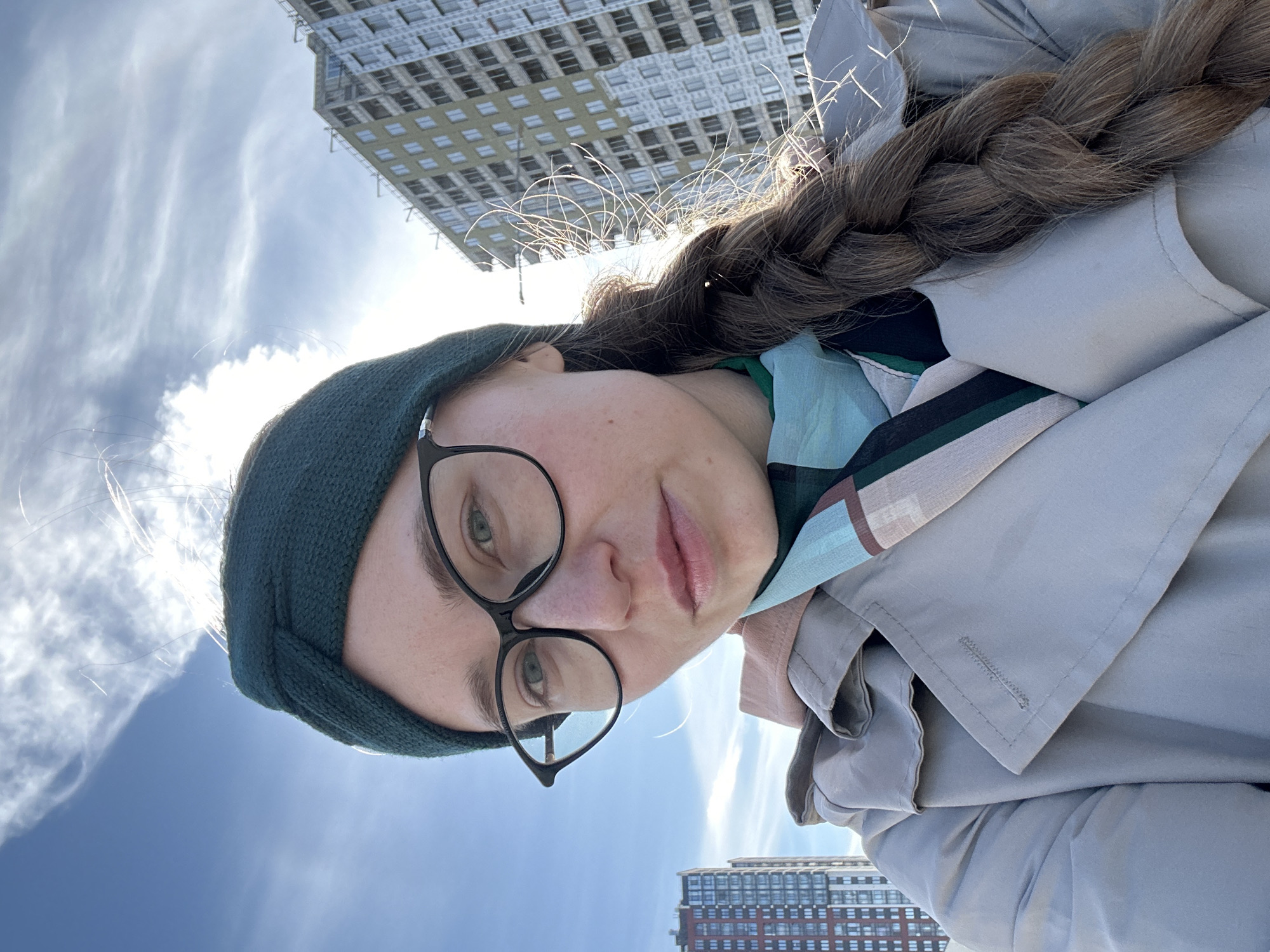A selfie of a woman wearing a green knit headband and black framed glasses, a light grey trench coat and a blue, green and pink patterned scarf. Her hair is light brown and braided. The background features a partly cloudy, bright blue sky. Behind the woman are two buildings. The buildings and the style of the buildings indicate that the location is Saint Petersburg, Russia.