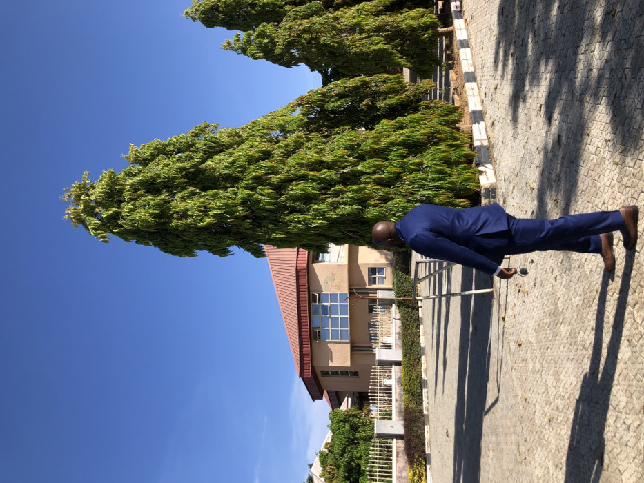 An individual in a blue suit and brown shoes poses outdoors, holding a pair of sunglasses. The location appears to be a paved area with trees, a building with a red roof, and a blue sky in the background. The image is oriented sideways, requiring a 90-degree rotation to view it upright. The lighting is bright, casting shadows on the paved surface.