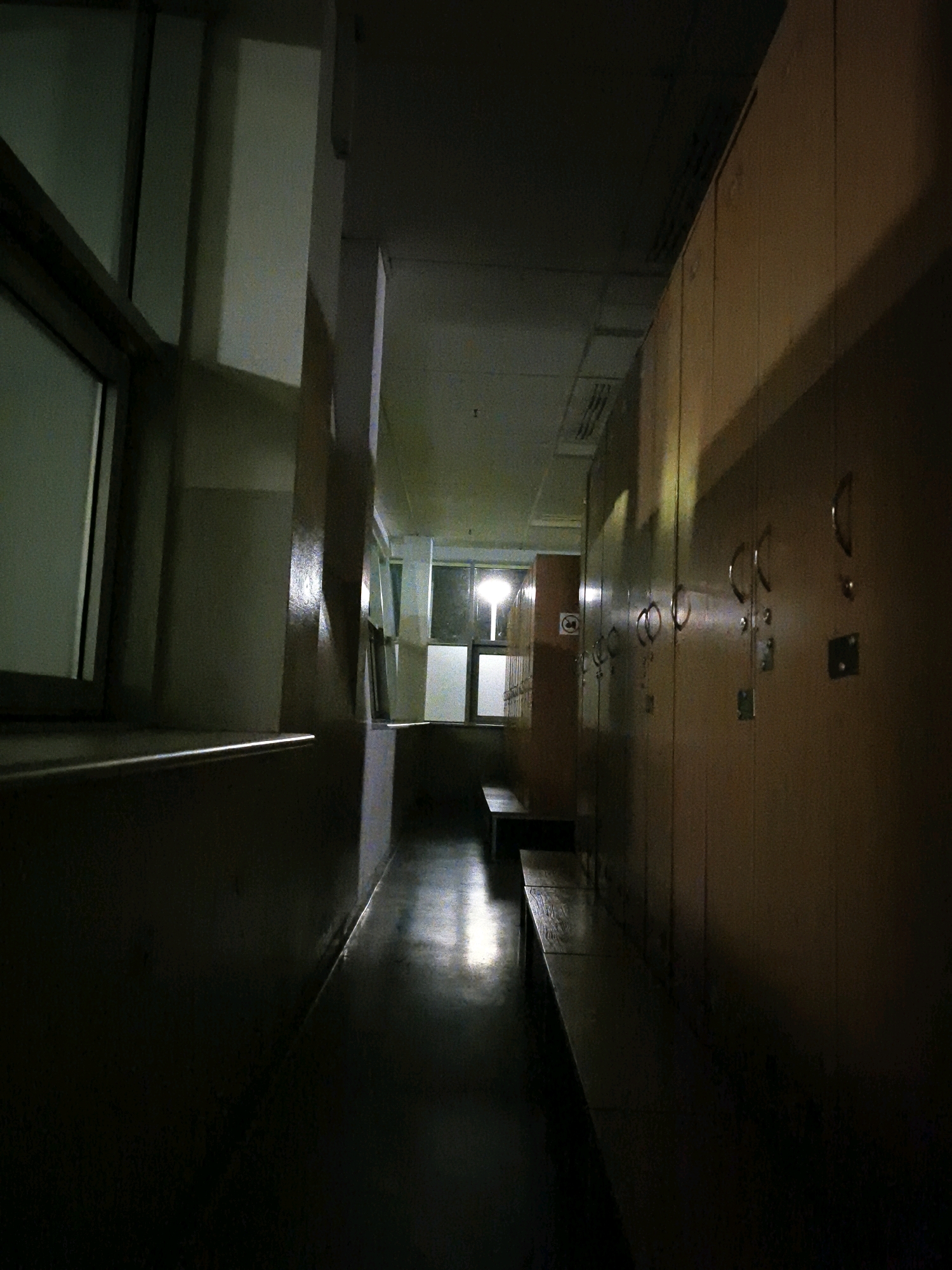 The image depicts a dimly lit hallway, likely within an institutional setting such as a school or locker room, as evidenced by rows of lockers on the right side of the frame. A long bench runs along the left side of the hallway, parallel to the lockers. The floor is dark and appears to be polished, reflecting the faint light that enters from a window at the far end of the hallway. The window at the end of the hallway is illuminated from within, suggesting a light source on the other side. The overall atmosphere is one of quiet emptiness, with no people visible in the scene. The style of the lockers and the hallway suggest a mid-20th-century construction. Without further information or context, it is impossible to determine the specific location within the city of Tczew, Poland.