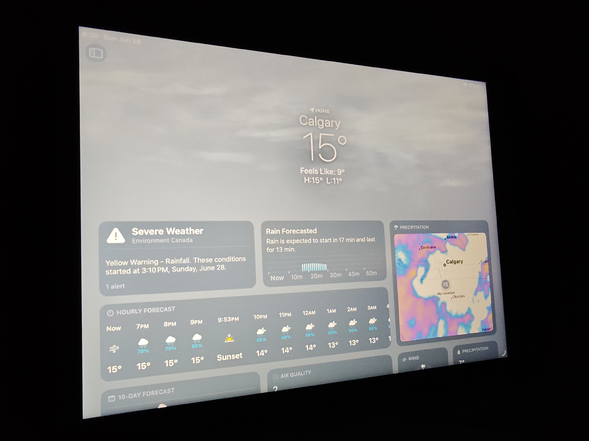 The image shows a tablet displaying the weather application for Calgary, Canada. The time is 6:30 AM on Sunday, June 28th. The current temperature in Calgary is 15 degrees Celsius, with a "feels like" temperature of 9 degrees Celsius. The high for the day is 15 degrees Celsius and the low is 11 degrees Celsius. A severe weather alert for rainfall is active, with a yellow warning issued by Environment Canada. The rain is expected to start in 17 minutes and last for 13 minutes. The hourly forecast shows cloudy conditions with a 70% chance of precipitation for the next few hours, with temperatures remaining around 15 degrees Celsius. The 10-day forecast is also visible. A precipitation map of the Calgary region is displayed, showing areas of light to moderate rainfall. The tablet is displaying a cloudy sky background.