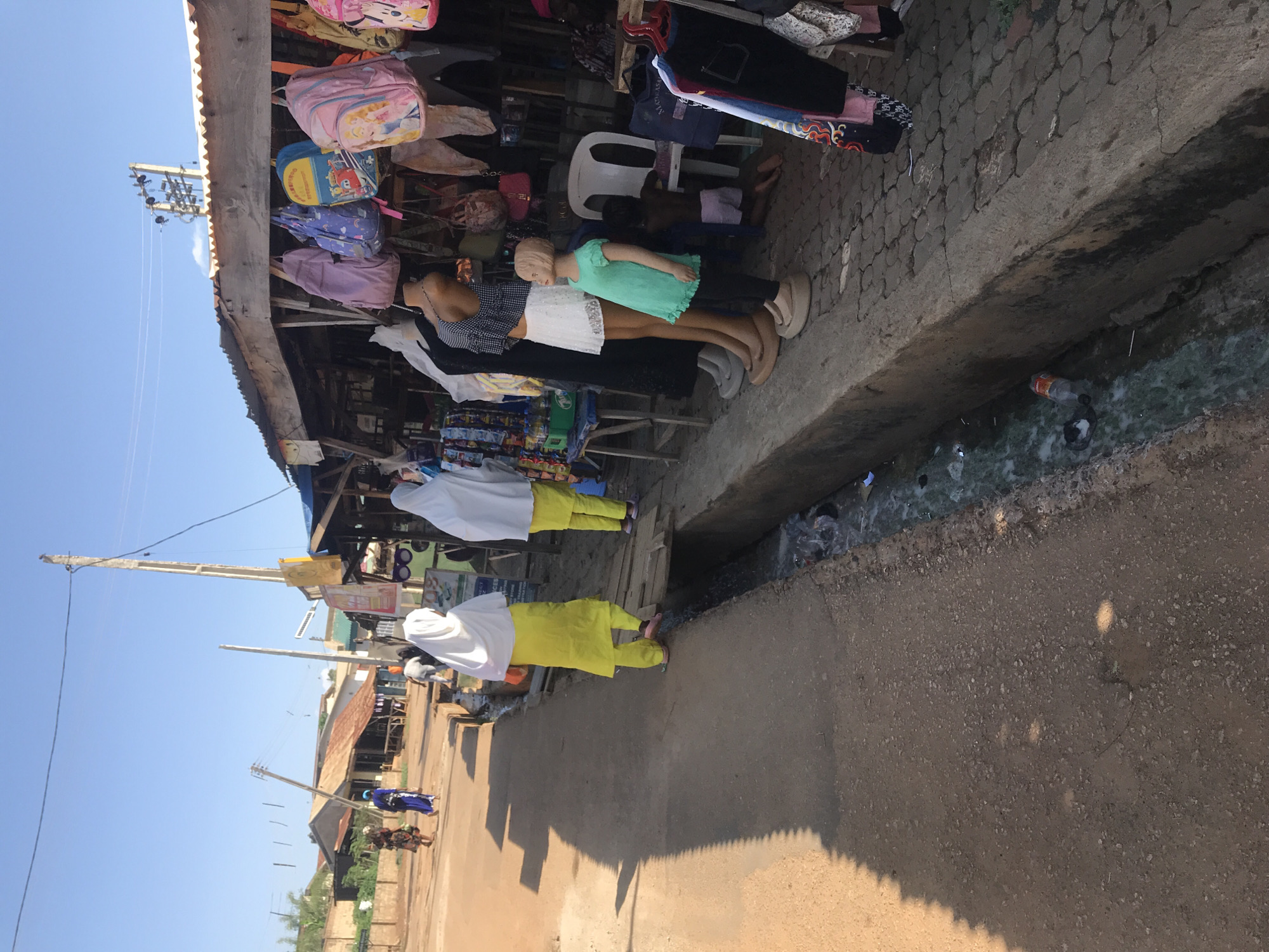 This street-level photograph, taken in Mandate Estate, Ilorin, Nigeria, captures a vibrant outdoor market scene under a clear, bright blue sky. The image appears to be rotated, showing the horizon vertically.
In the foreground and midground, a bustling market stall constructed from wood and corrugated iron is visible. It is heavily laden with merchandise, including numerous colorful backpacks, some featuring cartoon characters, and various items of clothing hanging for display. Several mannequins are prominently featured, showcasing apparel such as dresses, skirts, and shirts; one is a child-sized mannequin wearing a teal top. A small child is seated on a white plastic chair behind the mannequins.
The market stall sits alongside a concrete walkway with circular textured patterns. Immediately next to the walkway is an open ditch containing murky water and scattered litter, including plastic bottles and bags. Beyond the ditch lies a unpaved, dusty road. Strong, sharply defined shadows stretch across the road and walkway, indicating bright sunlight, characteristic of midday or early afternoon.
On the left side of the road, two individuals wearing white head coverings and distinctive bright yellow long-sleeved outfits are visible, either standing or walking. Further down the road, more people and buildings are discernible in the distance. Utility poles with numerous power lines crisscross the sky, adding to the urban landscape. The scene conveys a busy, functional market environment. No clearly legible text is visible on any signs or merchandise.