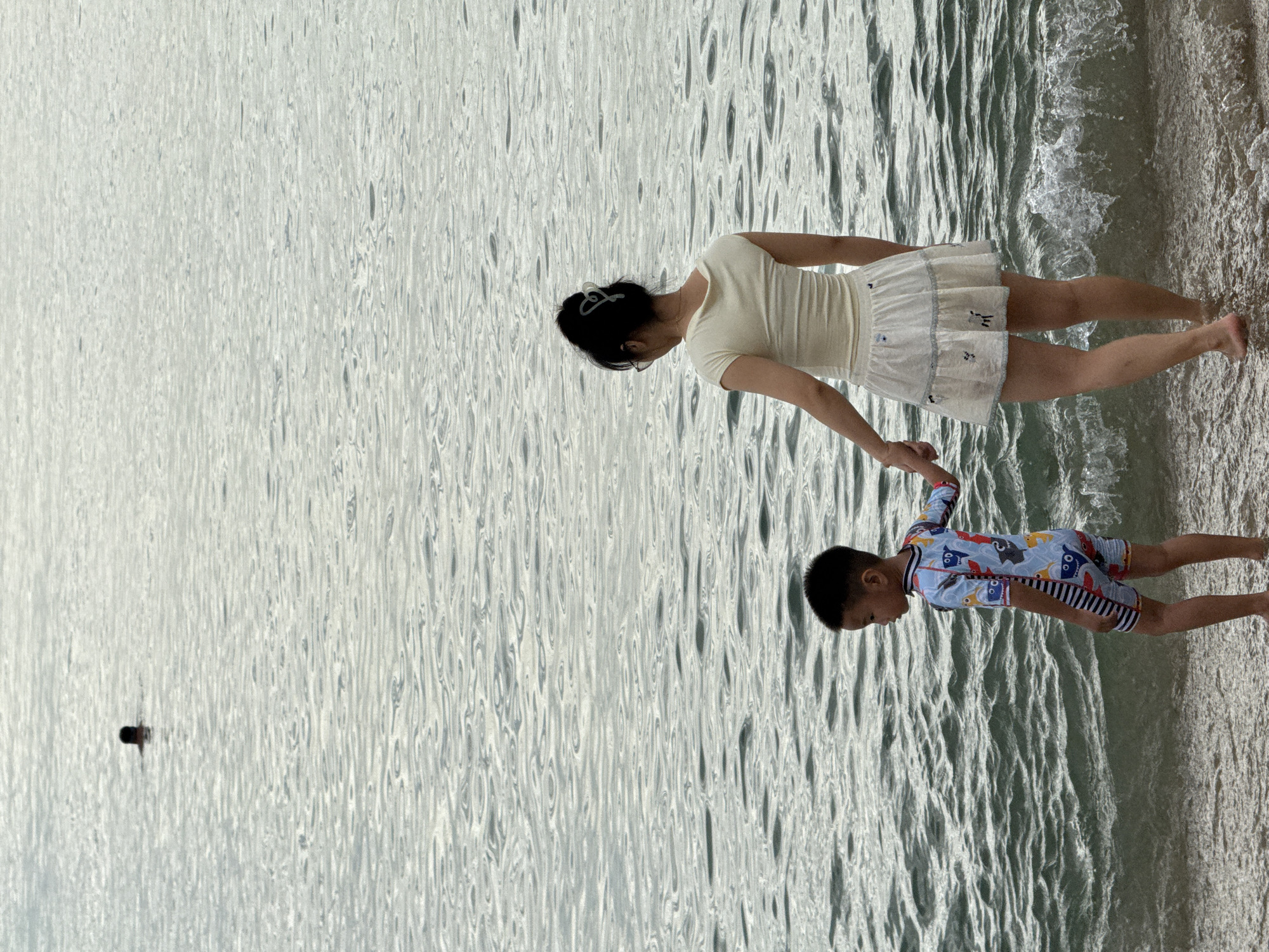 Two people, a child and an adult, stand on the sandy edge of the ocean. The child, wearing a blue swimsuit with a pattern of sea creatures, holds the adult's hand. The adult, facing away from the camera, wears a pale yellow top and a white skirt. The water is calm with gentle waves lapping the shore. In the distance, another person is visible swimming. The scene appears to be set on a beach in Nha Trang, Vietnam.