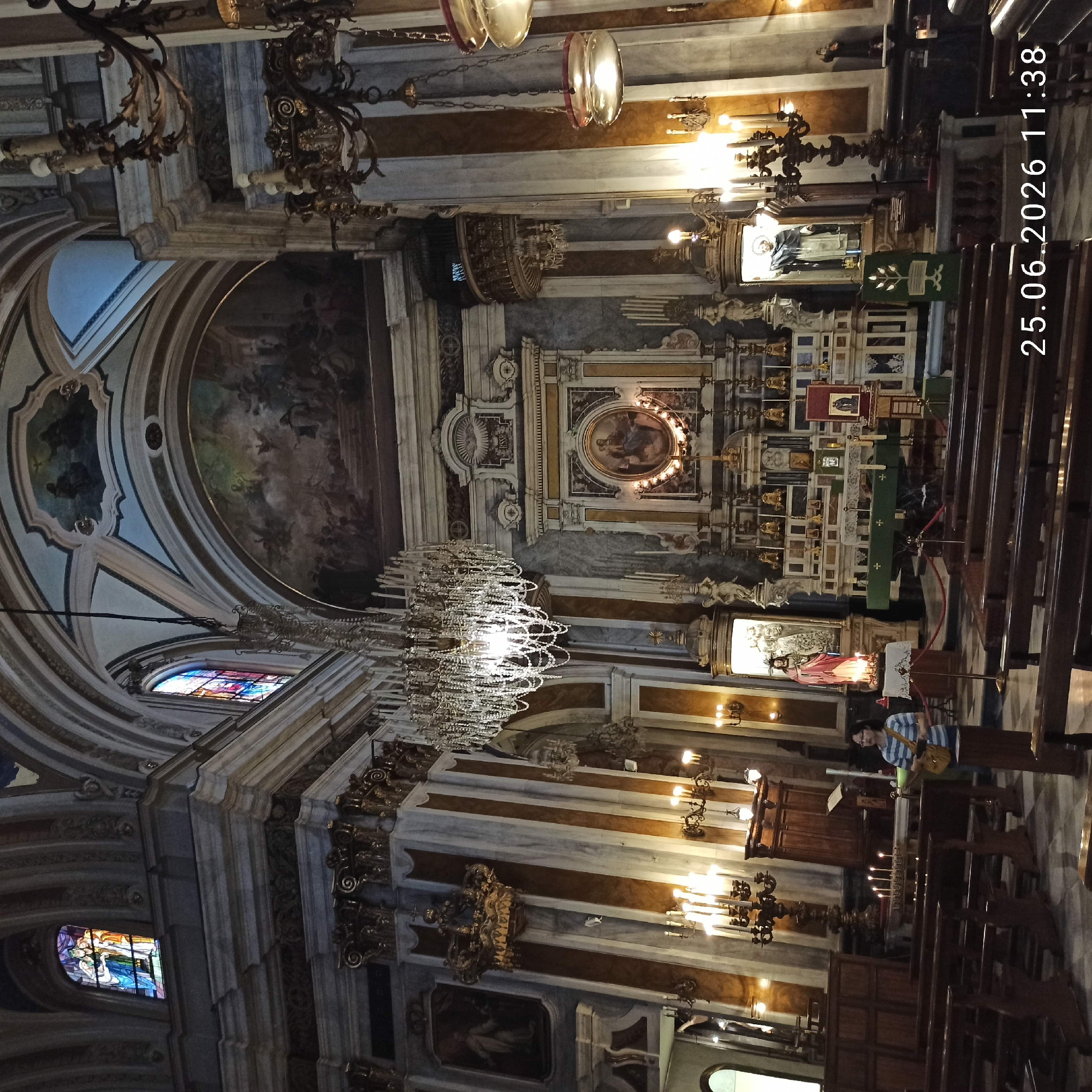 This is a vertical, indoor photograph of a church interior, bathed in artificial light. The scene is dominated by ornate architecture, including arches, columns, and decorative moldings. A large, crystal chandelier hangs from the ceiling, sparkling with light. Several stained-glass windows with vibrant colors are visible, casting colorful patterns on the walls.
In the background, an altar is adorned with religious iconography, including statues and paintings. Several rows of wooden pews fill the main area of the church, suggesting a place of worship. The overall impression is one of grandeur and solemnity, typical of historical religious buildings.
A person is visible on the right side of the image, standing near the pews and looking towards the altar. They are wearing a striped shirt and carrying a bag. Another person is partially visible in the lower left corner. The time stamp "25.06.2026 11:38" is overlaid on the image, indicating the date and time the photo was taken. There are no discernible weather cues, as the photograph is taken indoors. The location context is unknown.