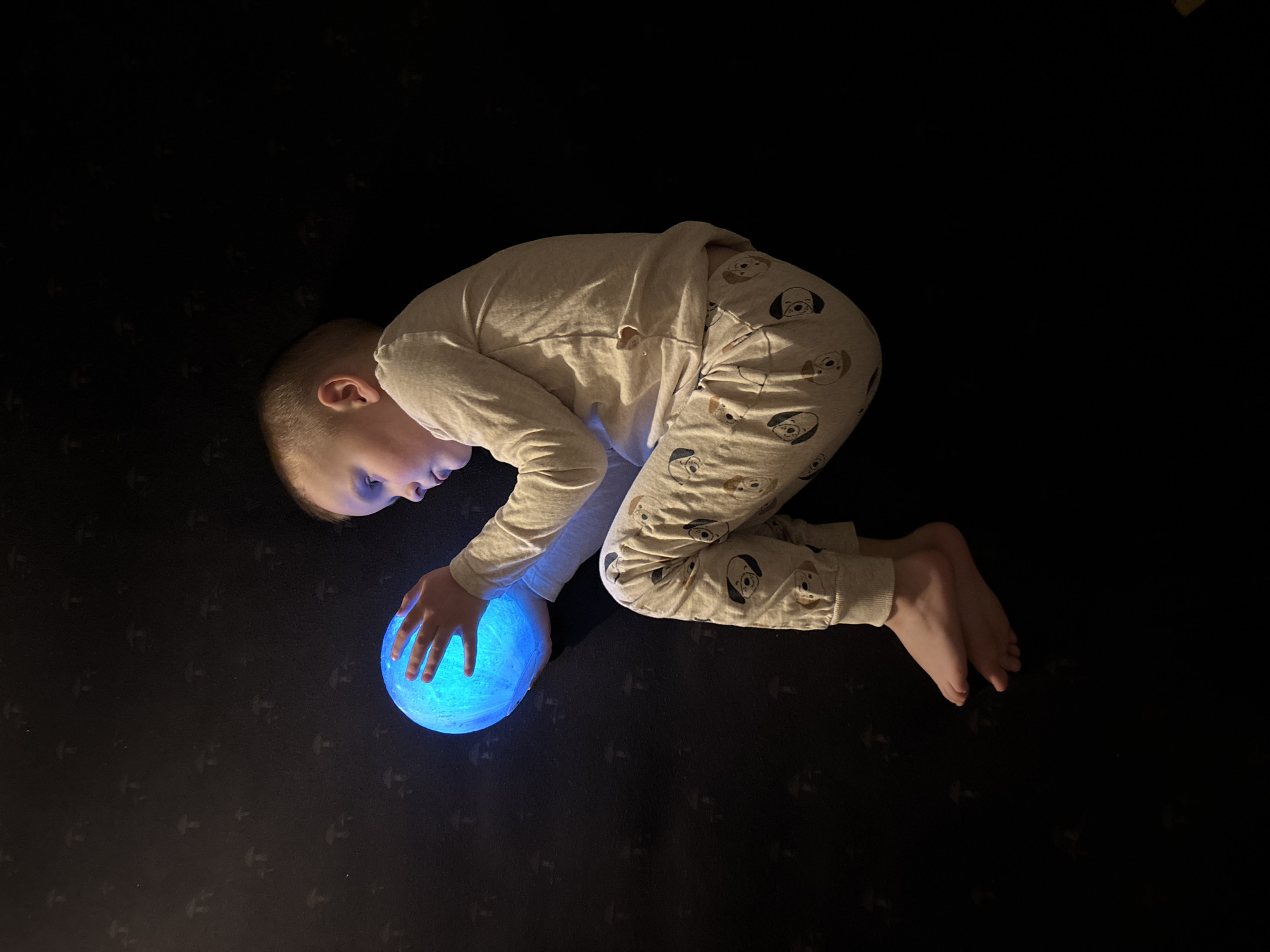 This image depicts a young child, likely a male, with light-colored hair, situated in a dimly lit indoor environment within the city of Chaikovsky, Russia. The child is positioned in a curled or seated posture, facing towards the right side of the frame, with knees drawn upward. He is wearing a light-colored, long-sleeved pajama set consisting of a plain top and pants adorned with a repeating pattern of cartoon dog faces. Both of the child's bare hands are clasping a spherical object, approximately the size of a small melon, which emits a vivid, glowing blue light. This light serves as the primary illumination source, casting a blue hue onto the child's face, hands, and the immediate surroundings. The background is a dark surface featuring a subtle, repeating pattern of small, darker mushroom-like silhouettes. The child's gaze is directed downward and forward, focused on the glowing object he holds.