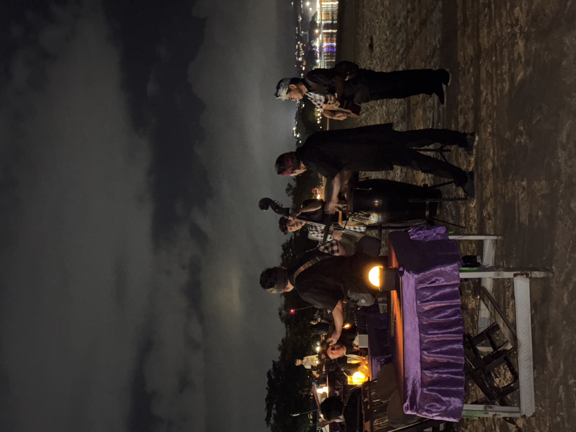 This is a nighttime photograph taken on a beach in Kuta Selatan, Indonesia. The scene depicts a band performing for diners at tables set up on the sand. A group of four musicians is visible, playing a double bass, conga drums, and a guitar. They are all wearing dark clothing and headbands. In the foreground, a table is covered with a purple cloth, and a lit lantern sits on it. Several other tables are visible in the background, with diners seated and illuminated by lanterns. The ocean is to the right, with distant city lights visible across the water. The sky is dark with some visible clouds.