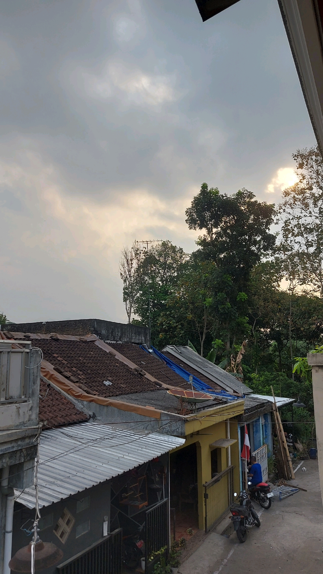 This image depicts a residential area with a view of rooftops and trees under a cloudy sky. The architecture suggests a tropical or Southeast Asian setting, likely in Salatiga, Indonesia, given the hints of a flag and dense foliage.
In the foreground, multiple rooftops are visible, some covered with terracotta tiles and others with corrugated metal. One roof is partially covered with a blue tarp. A satellite dish is mounted on one of the tiled roofs. Adjacent to the rooftops, a narrow alleyway runs alongside a yellow building. Two motorcycles are parked in the alley. A person in a blue shirt is standing near the entrance of the yellow building, interacting with another person who is seated. The entrance is adorned with a red and white flag, likely the Indonesian flag.
In the background, dense green trees create a natural backdrop. The sky is overcast with breaks of sunlight peeking through, suggesting it might be late afternoon or early morning. The overall atmosphere is one of a typical urban or semi-urban neighborhood in Indonesia.