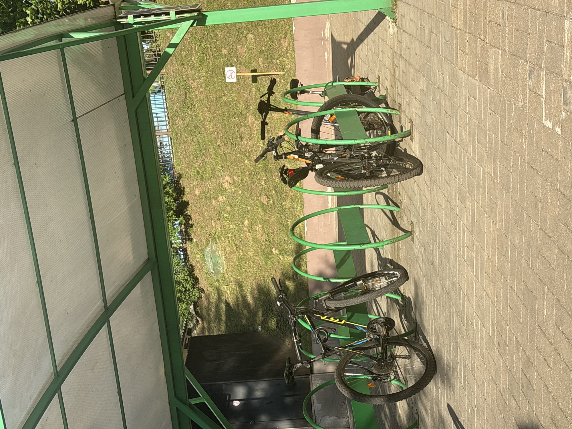 The image depicts a bike rack at what appears to be a covered bike parking area. Two bicycles are secured in the green, circular bike rack. The bikes are of different brands, one with black and yellow trim. The area around the rack is paved with light-colored bricks, and there is a grassy area nearby. A sign prohibiting something (likely cycling) is present in the background. It is daytime with ample natural lighting, casting shadows on the pavement. There is no evidence of human presence. The location appears to be in Minsk, Belarus.
