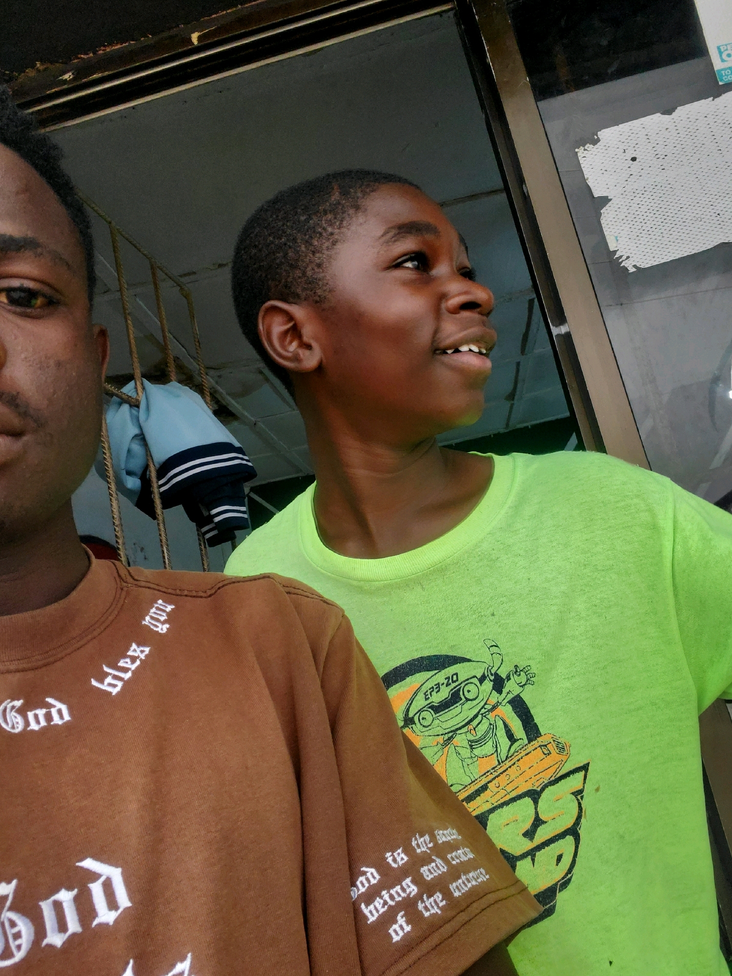 Two young Black males are shown from the chest up, posed in front of a doorway or window. The male on the left is partially visible, with only his face, left shoulder, and upper chest in frame. He has dark skin, short black hair, and is wearing a brown long-sleeved shirt with white gothic-style lettering that says "God bles you". The male on the right is more fully visible from the shoulders up. He has darker skin and short black hair, and is wearing a neon green t-shirt with a graphic of a cartoon robot holding a yellow object. Both subjects appear to be looking slightly off-camera to their right. The background includes a metal grate, a light blue jacket with dark stripes hanging on the grate, and a window with a peeling white sticker on it. The scene appears to be outdoors or in an open-air structure. The lighting suggests it is daytime. The location is identified as Jos, Nigeria.