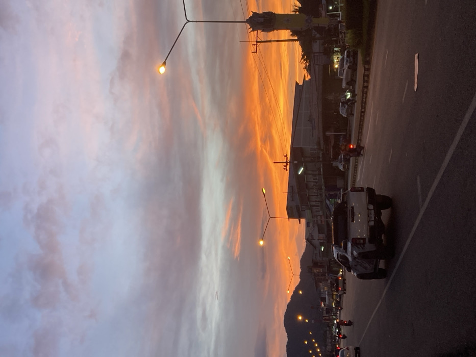 The image depicts an evening street scene in Bang Sawan, Thailand, characterized by a vivid sunset. The sky displays intense orange and red coloration near the horizon, gradually transitioning to purple and light blue with scattered clouds overhead. A multi-lane road, likely a main thoroughfare, stretches into the distance, with white lane markings visible.
In the foreground and mid-ground, a white pickup truck, possibly a Mitsubishi Triton, occupies the middle right lane, oriented away from the viewer. Multiple other vehicles, including passenger cars and several motorcycles, are present on the road, all appearing to move in the same direction. Some vehicles exhibit illuminated headlights and taillights. Numerous streetlights are active, emitting a warm yellow light that reflects subtly on the road surface.
Along the right side of the road, a series of low-rise commercial or mixed-use buildings line the street. Utility poles and overhead power lines are visible on this side. A tall, ornate yellow-gold structure or monument stands prominently on the far right. In the distant left, a silhouette of hills or mountains is visible against the twilight sky.