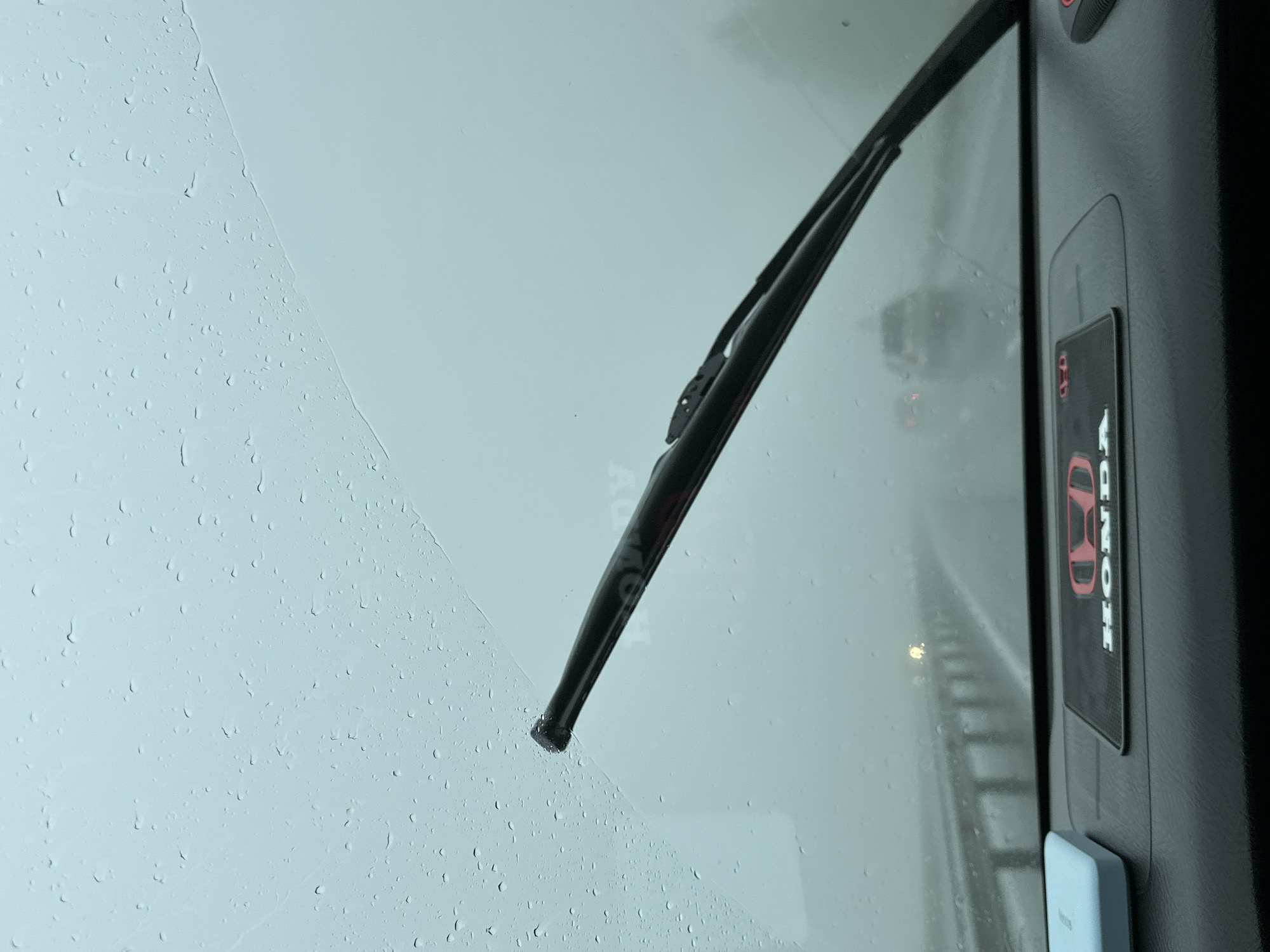 The image shows a view from inside a vehicle driving on a highway in the city of Pionersky, Russia. It is raining heavily, with numerous raindrops visible on the windshield. A windshield wiper is positioned across the glass, partially obscuring the view of the road ahead. In the distance, other vehicles are visible on the highway, their taillights faintly glowing through the fog and rain. The road is wet and appears to be a multi-lane highway with a metal guardrail on the left side. The interior of the car is visible in the foreground, with a black dashboard and a blue object on the left side. A black mat with a red Honda logo is placed on the dashboard. The overall atmosphere is hazy and obscured due to the inclement weather.