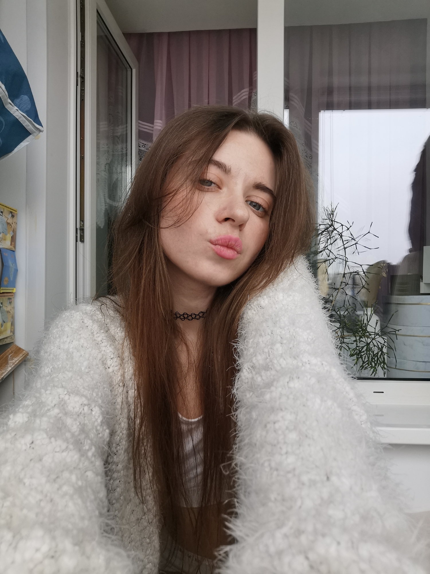 A young woman with long brown hair is positioned in the foreground, facing the camera with a slight tilt. She has light skin, blue eyes, and is pursing her lips in a kiss gesture. She is wearing a fuzzy white sweater and a black choker necklace.
Behind the woman, there's a window with a white frame. Visible through the window are blurred, out-of-focus shapes and a bare-branched plant in a pot. To the left of the window is a section of white wall with visible items, including a blue bag with white patterns and a box decorated with images. A second window is on the left. The location is indoors. The city is Unknown, Unknown.