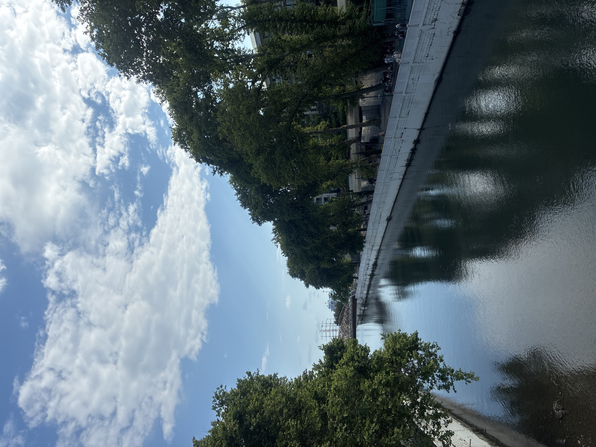 The image depicts a canal lined with trees and buildings. On the left, a clear blue sky is adorned with fluffy white clouds. The canal's dark water reflects the sky and surrounding trees. Along the canal's edge, a stone embankment is visible, with trees providing shade. In the distance, a bridge can be seen. The scene suggests a leisurely day, possibly a weekend, with people gathered along the canal bank, some sitting and others walking. The atmosphere appears calm and relaxed, with the dappled sunlight filtering through the leaves and reflecting on the water. The time of day seems to be daytime, given the bright sky and shadows. The location is identified as Sochi, Russia. There is no visible text in the image.