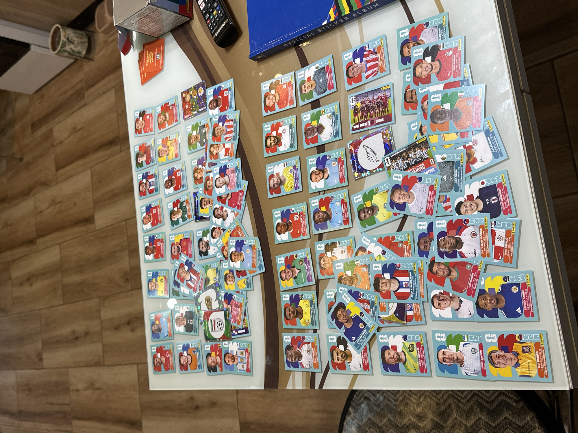 The image shows a collection of Panini FIFA World Cup Qatar 2022 stickers spread across a white table with a wood-patterned floor in the background. The stickers feature portraits of football players, along with their names, nationalities, clubs, and statistics. Some stickers are organized in rows and columns, while others are scattered. Several opened sticker packets are also visible, along with a blue official sticker album. The overall scene suggests a hobbyist organizing or displaying their sticker collection. The location is identified as Prisovo, Bulgaria.