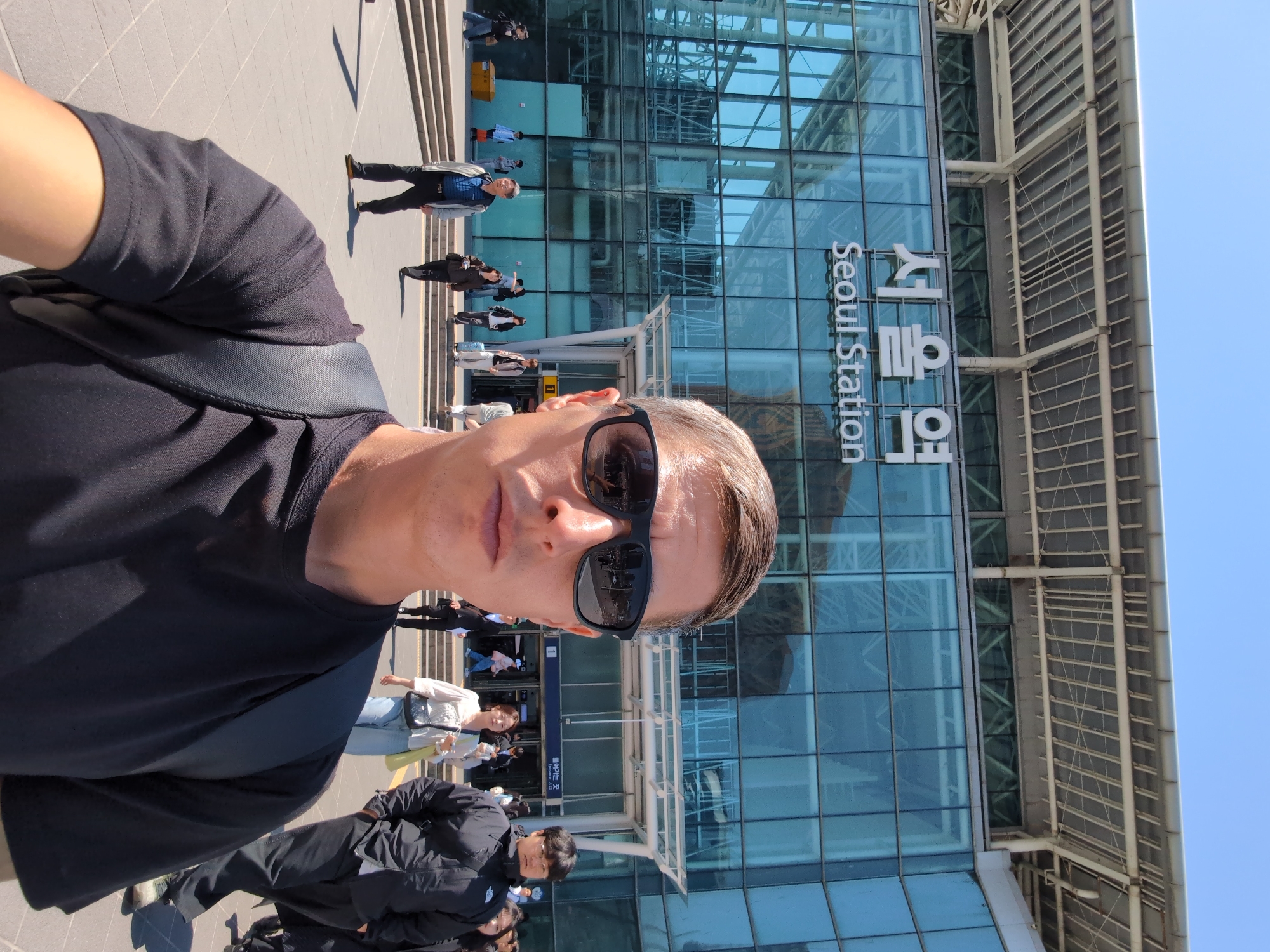 A medium shot of a person taking a selfie. The subject is a man with short, light-colored hair and sunglasses, wearing a black t-shirt with a black shoulder strap. He is outside Seoul Station, as seen from the large glass building bearing the words "Seoul Station" written vertically in English and Korean. People are visible entering and exiting the station. The weather is clear and sunny.