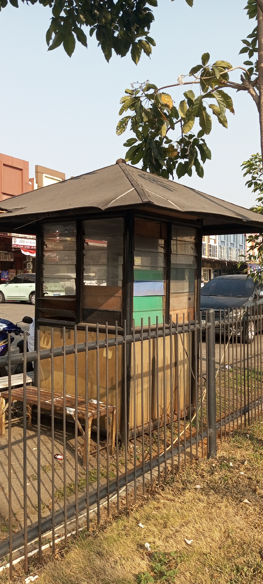 This image captures a street scene in Lambangjaya, Indonesia, focusing on a small, weathered guardhouse. The guardhouse, constructed with a dark, pitched roof and wooden panels, features several windows, some with horizontal louvers. Inside the windows, colored panels are visible, including green and blue. A metal fence, made of vertical bars, encloses the area in front of the guardhouse. A rusty metal bench sits behind the fence, partially visible. To the left of the guardhouse, a motorcycle with a person in a white shirt is parked. In the background, a light-colored car is on the road, and further back, buildings with windows can be seen. A black car is parked on the right side of the scene. Trees with green foliage and branches are present in the upper part of the image, with some leaves appearing dry. The ground in front of the fence is covered with dry grass. The sky is clear and light-colored.